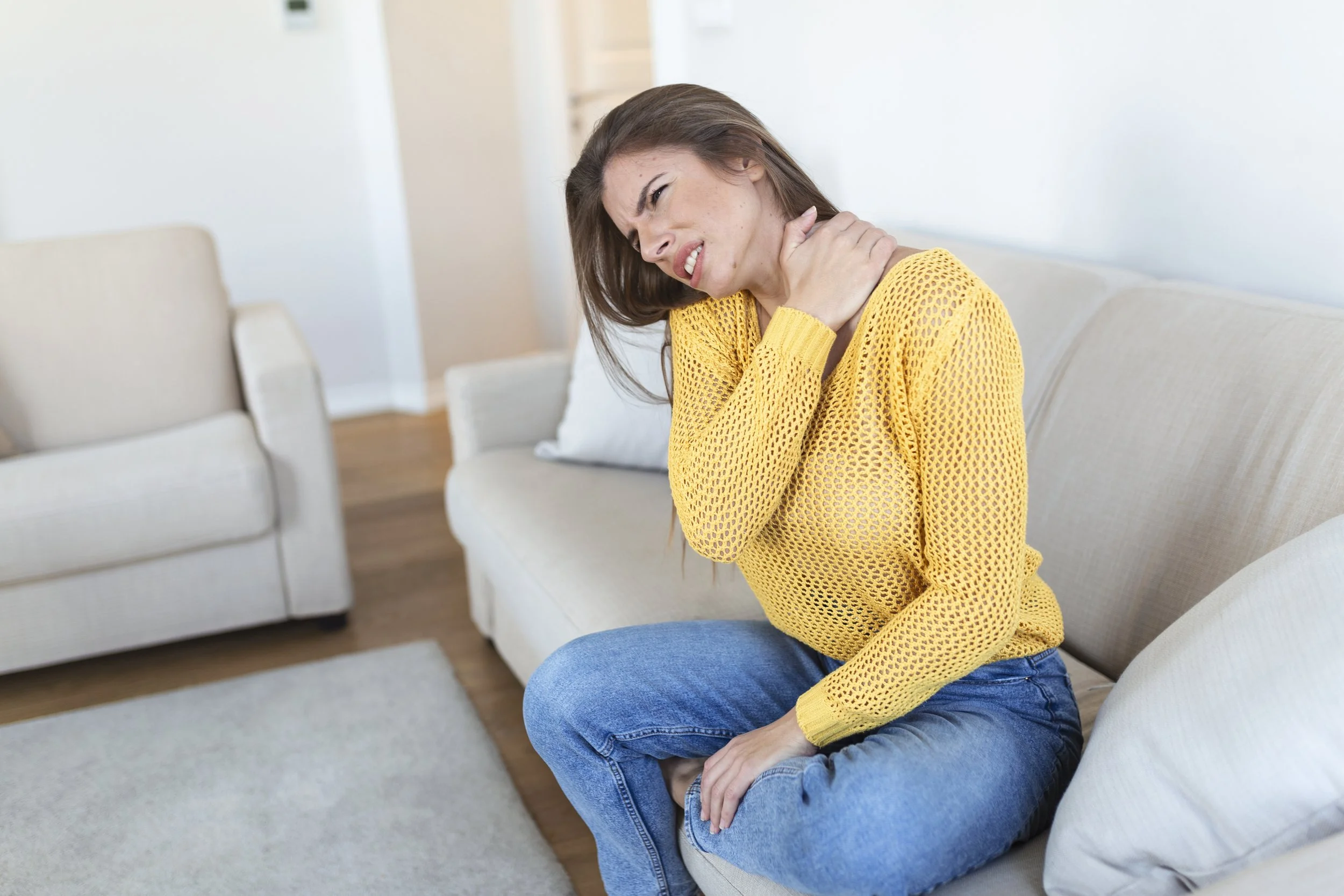 Woman sitting on a couch holding her neck and showing discomfort.
