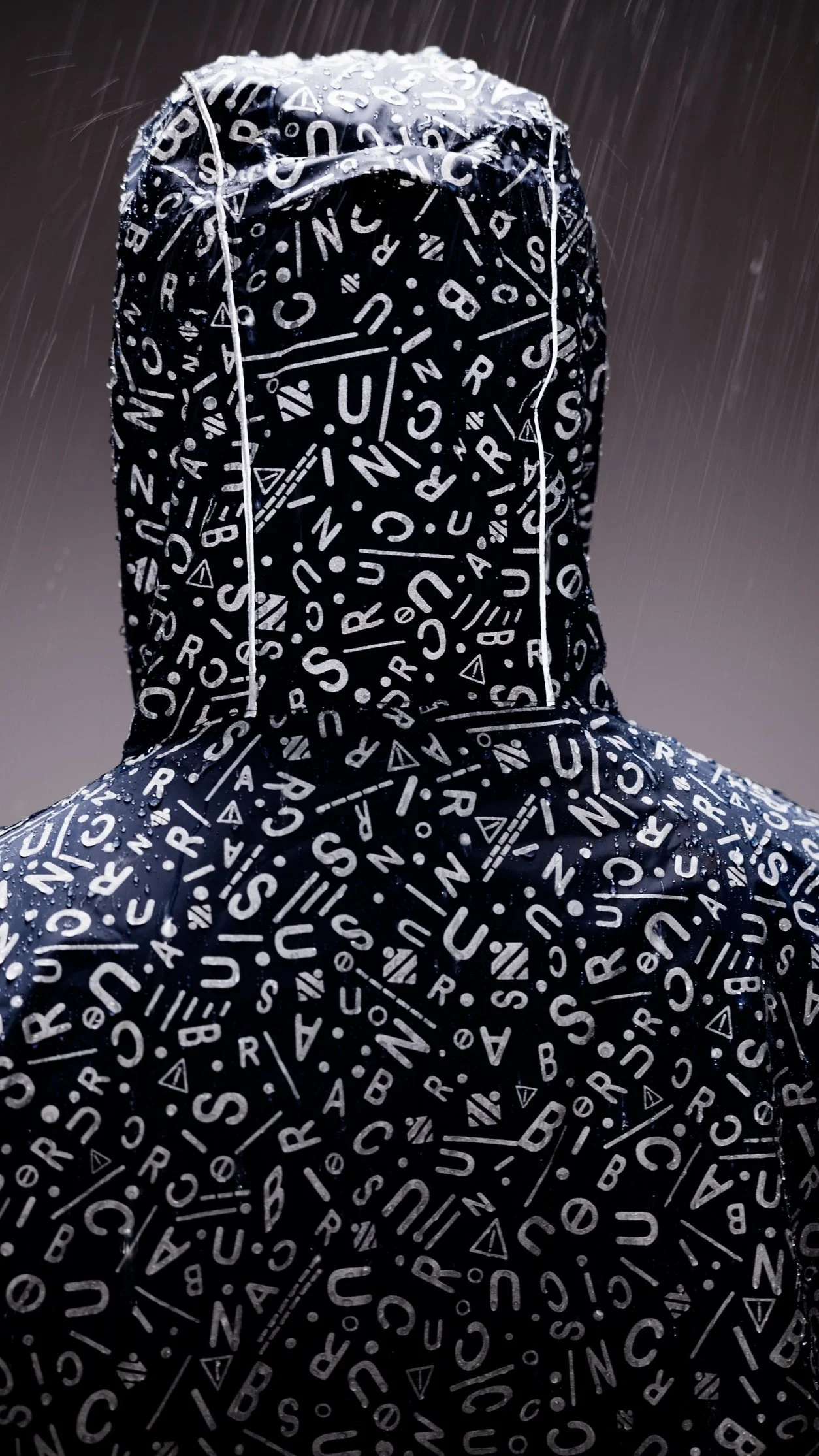 Homme vu de dos portant un manteau à capuche noir avec des lettres blanches et des symboles, sous la pluie.