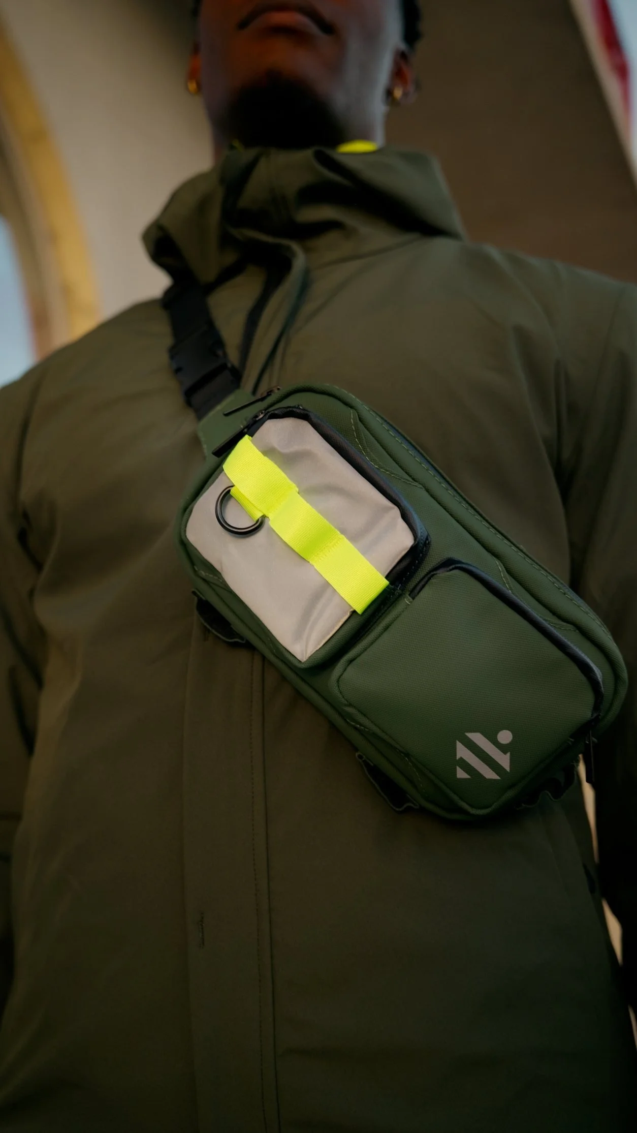 Homme portant une veste kaki et une ceinture banane verte avec un petit sac à l'avant, en intérieur.