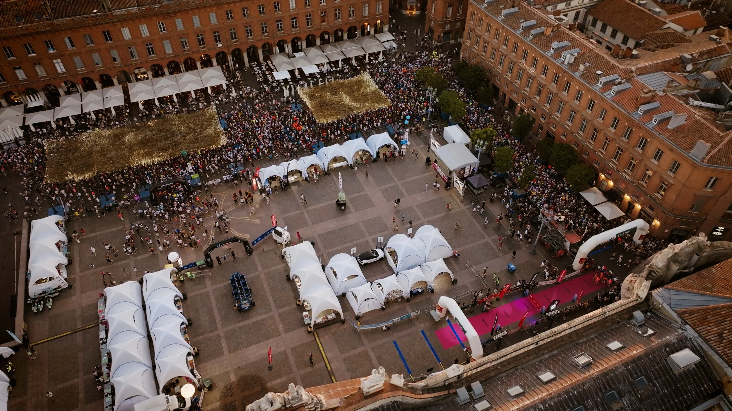Vue aérienne d'une grande place urbaine lors d'un événement, avec des tentes blanches, des zones pour la course avec un tapis rose, et une foule importante de personnes. Autour, des bâtiments anciens en brique, avec des toits en tuiles rouges, et des