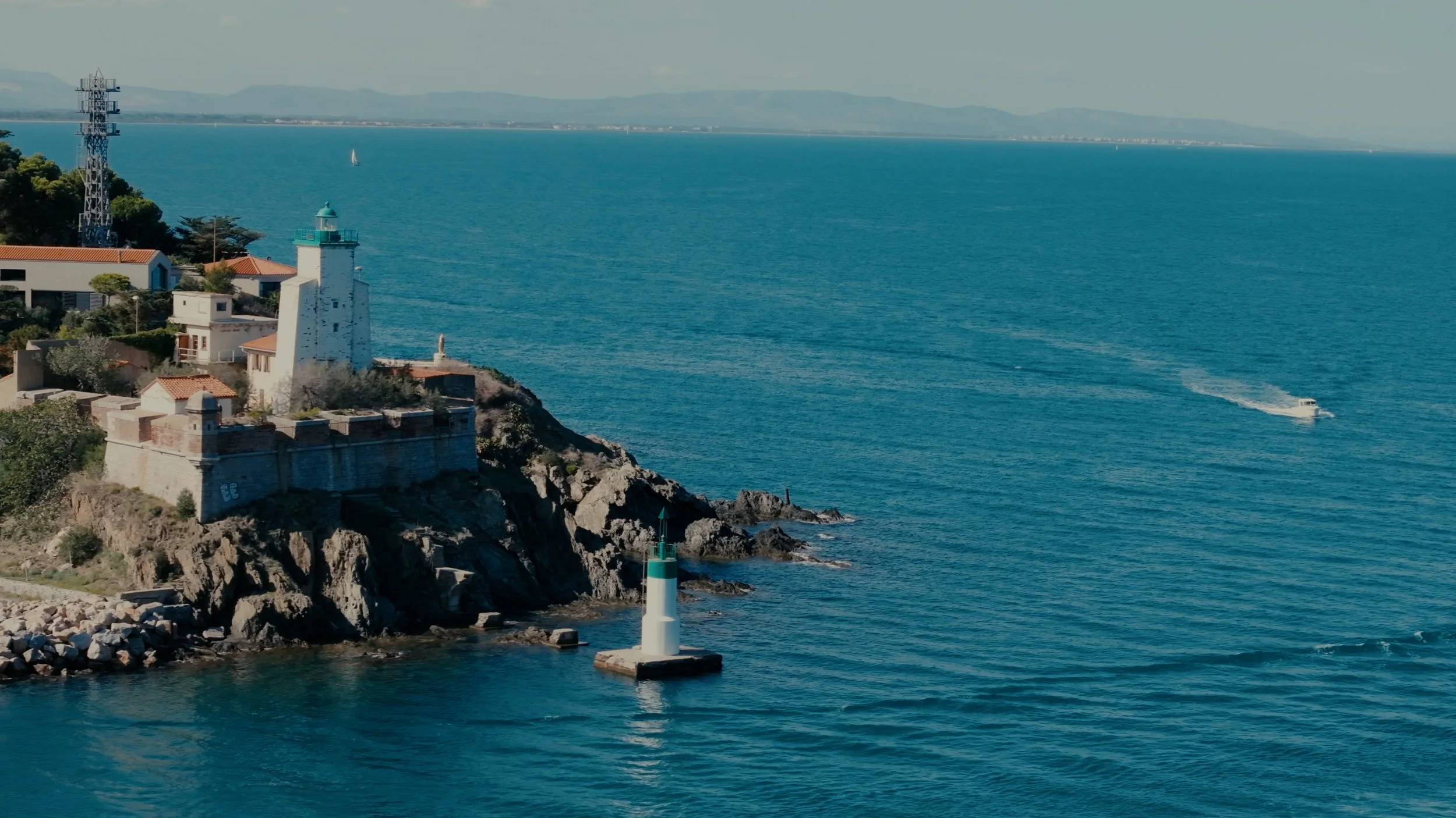 Phare blanc sur la mer avec des bâtiments et une maison sur un rocher, en arrière-plan des collines et un bateau à moteur dans l'eau.