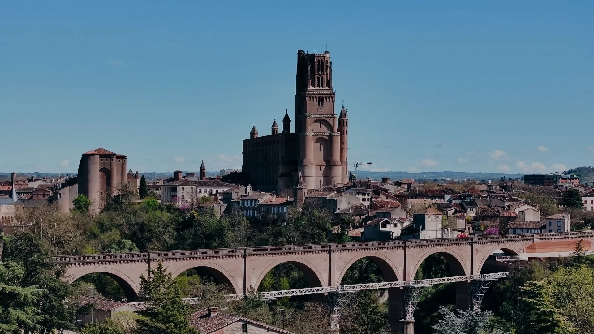 Une ville avec un pont en pierre traversant une vallée verdoyante et un château ancien en pierre avec une grande tour au centre, sous un ciel clair.