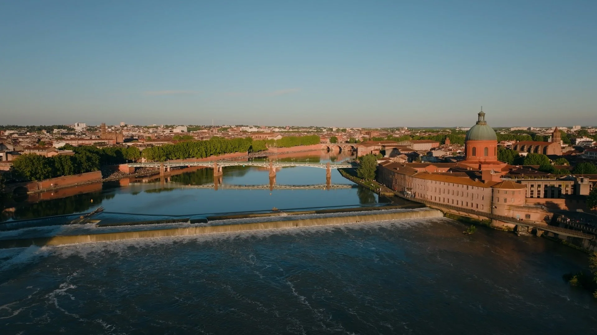 Vue d'une ville avec une rivière, plusieurs ponts, un bâtiment avec un grand dôme, arbres et bâtiments résidentiels en arrière-plan, sous un ciel clair.