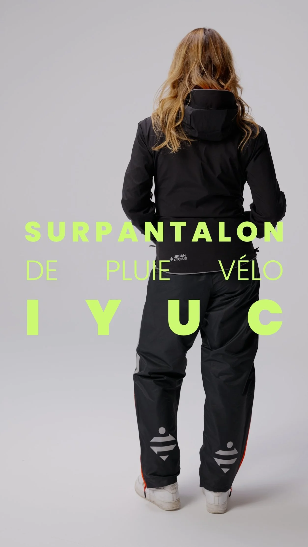 Une personne vue de dos porte un vêtement noir imperméable avec un capuchon sur fond gris.