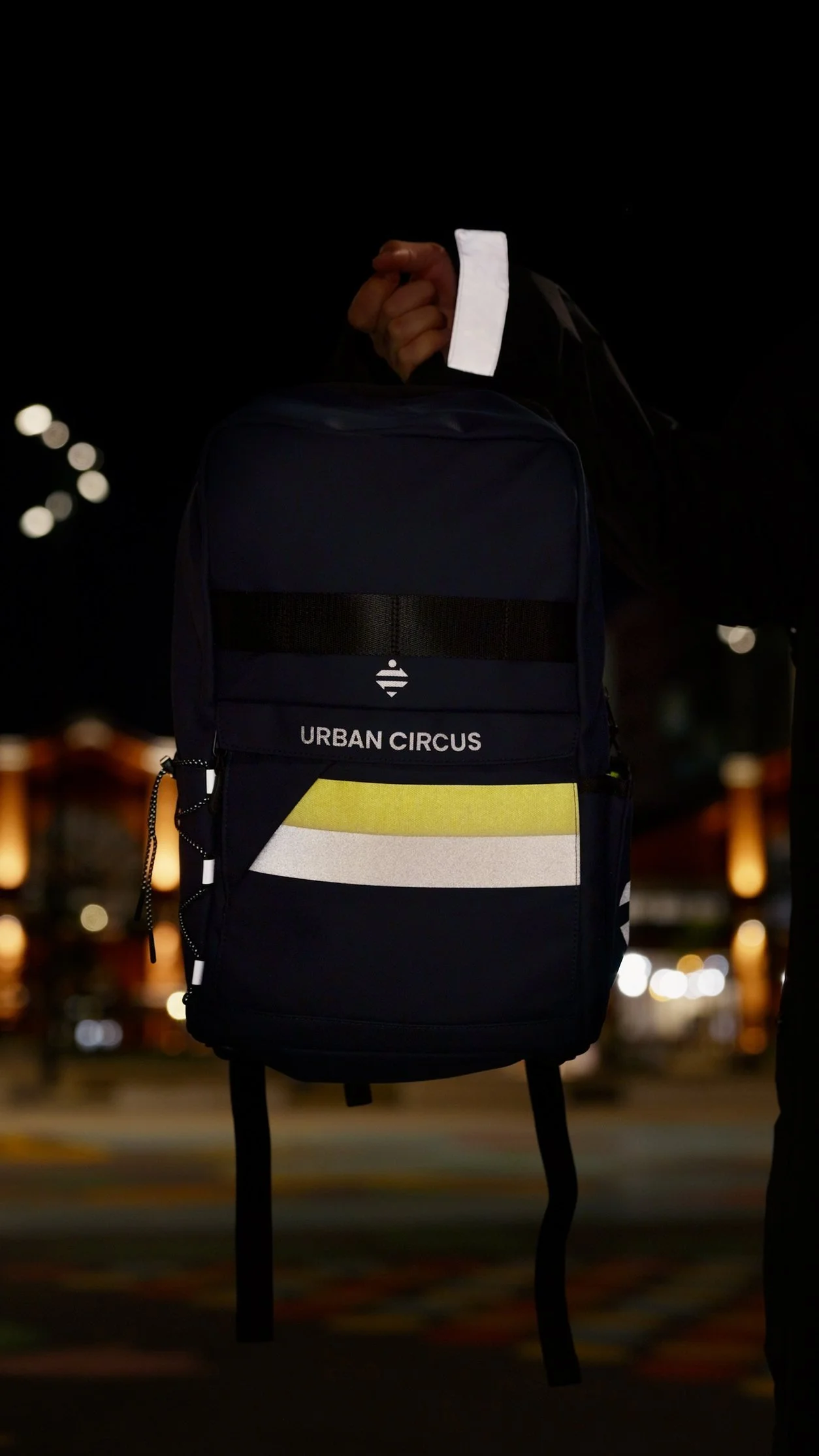 Un sac à dos noir avec la marque Urban Circus, porté par une personne dans un environnement nocturne. La personne porte une veste noire avec un contraste blanc, et la scène est éclairée par des lumières floues en arrière-plan.