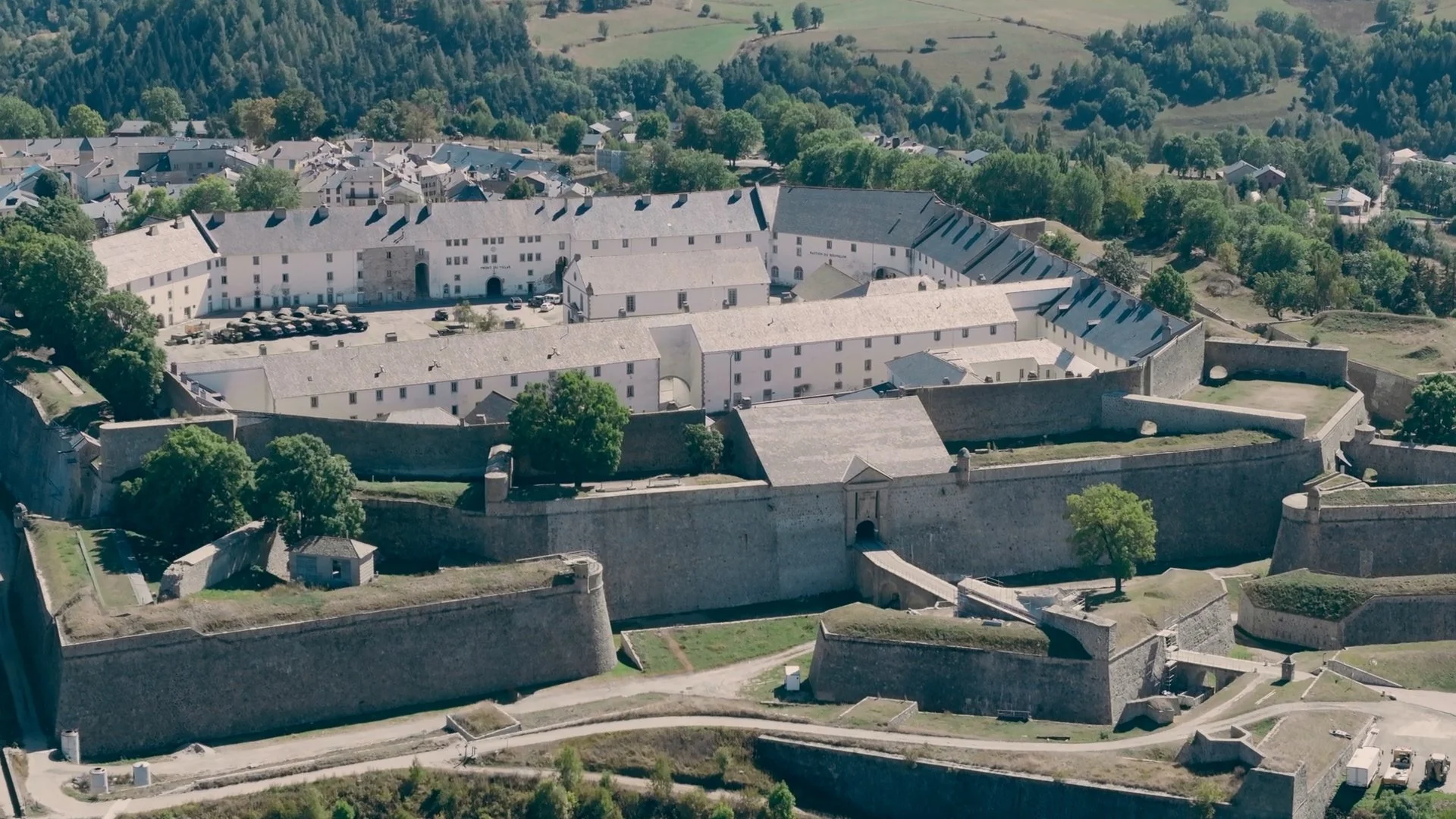 Vue aérienne d'une vieille ville fortifiée avec murs en pierre, bâtiments blancs et toits en ardoise, entourée de forêts et de collines verdoyantes, fortifications Vauban.