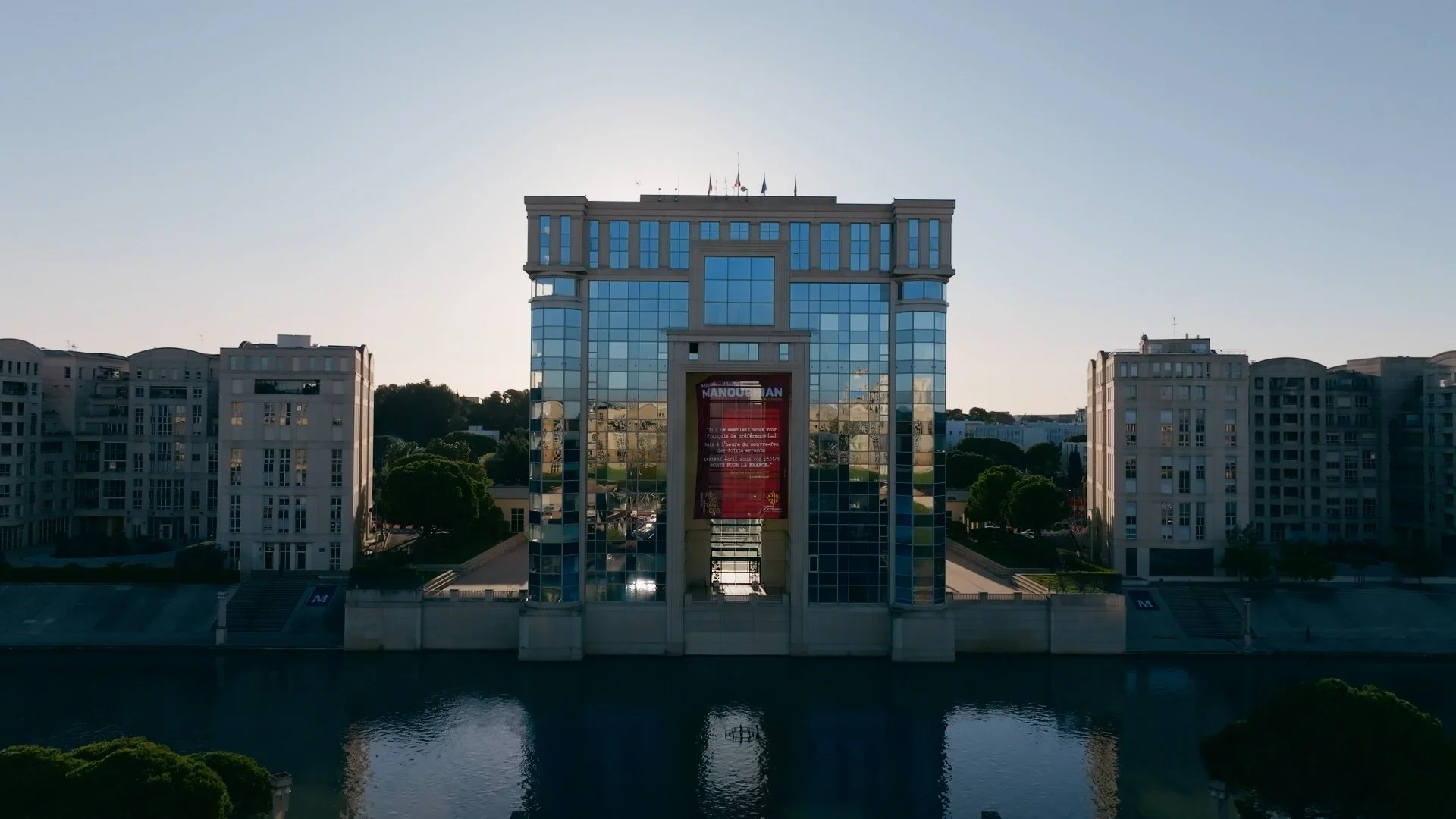 Tour d'affaires en verre avec bâtiments résidentiels environnants, reflet du soleil, et un panneau publicitaire en français sur la façade centrale. Région Occitanie