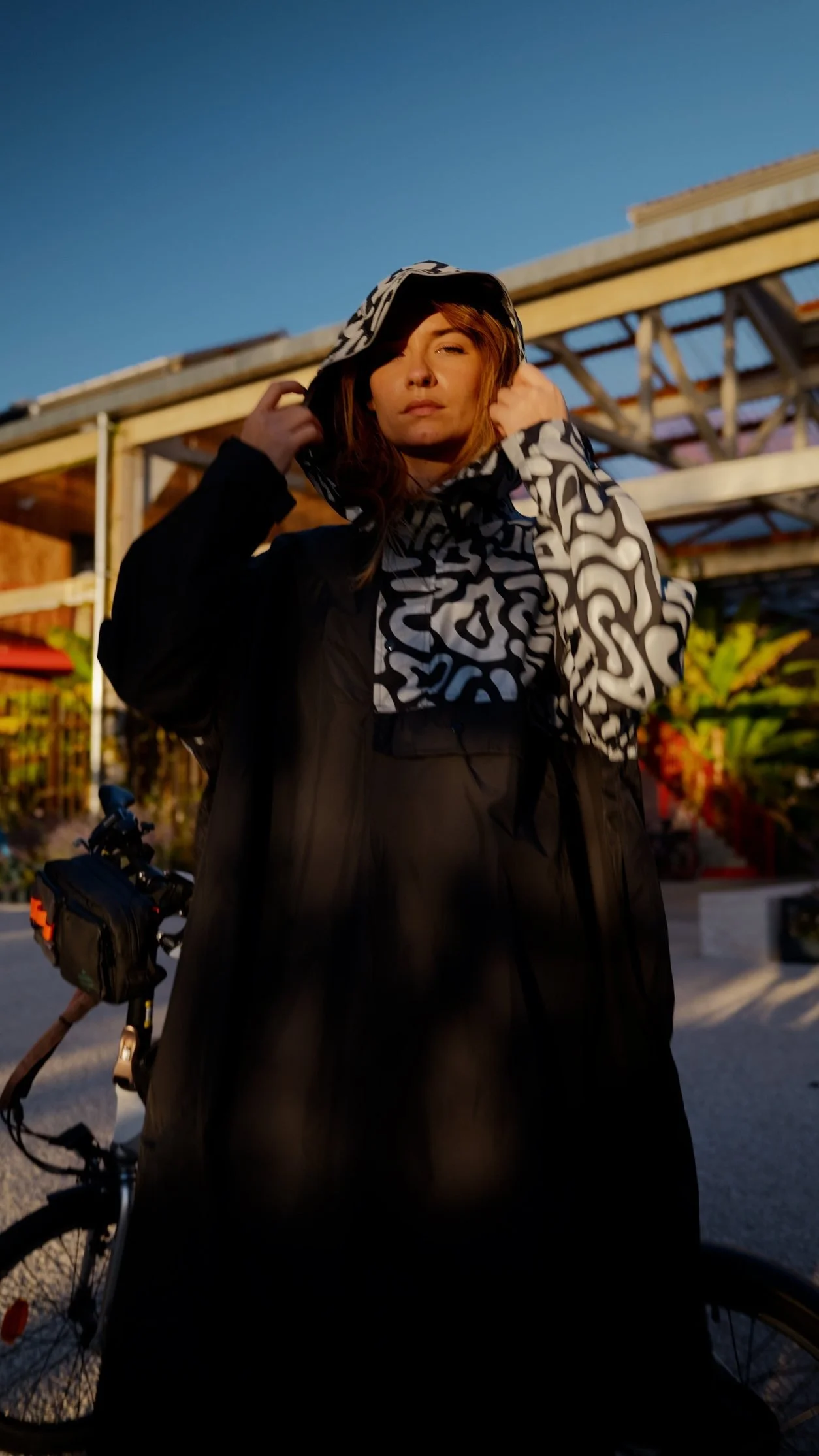 Femme portant une veste à capuche avec un motif noir et blanc, debout à l'extérieur avec un vélo, fond d'un ciel bleu au crépuscule.