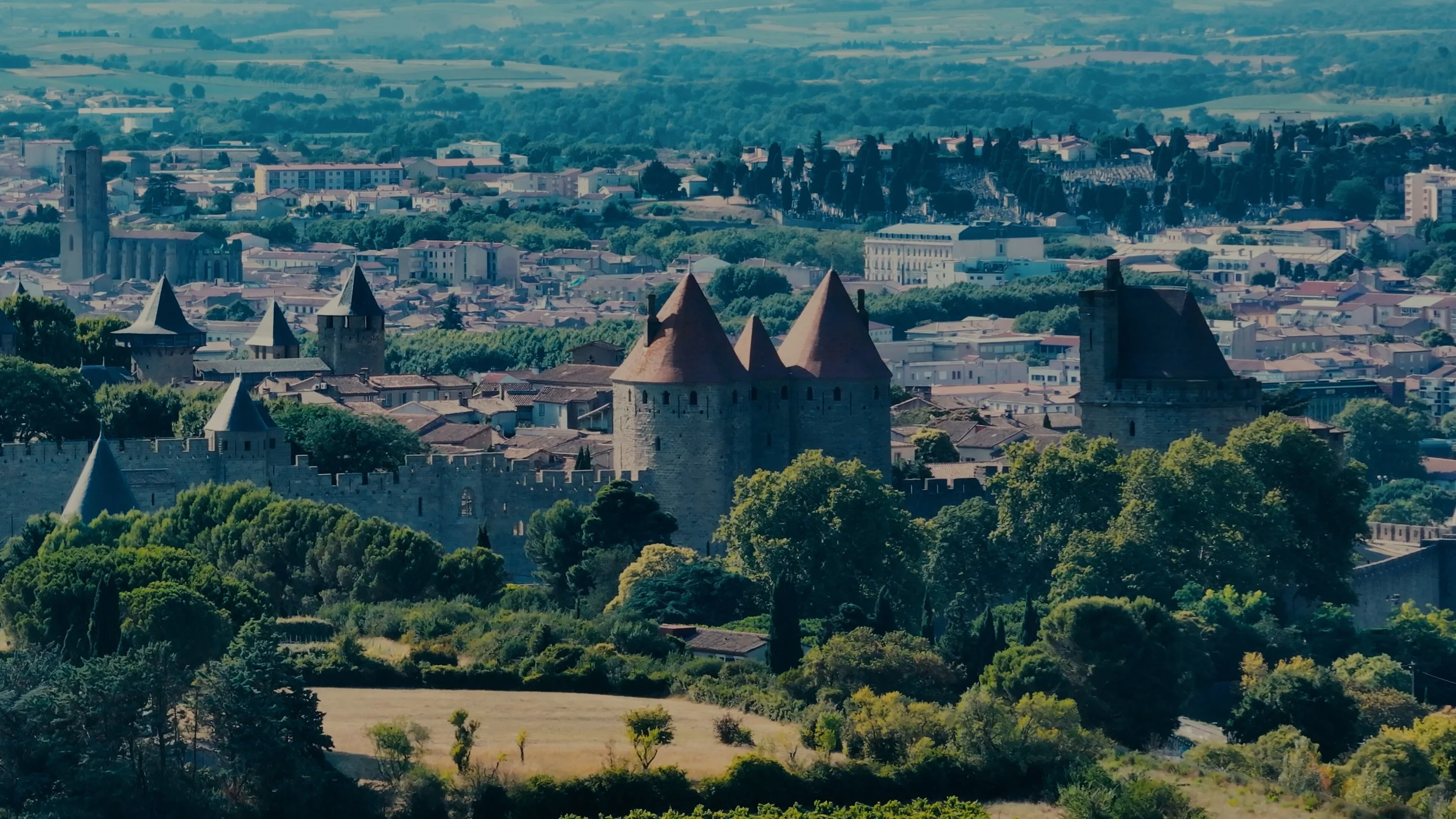 Vue panoramique d'une ville avec des tours médiévales en pierre et des toits en tuiles rouges, entourée de forêts et de collines à l'horizon.
