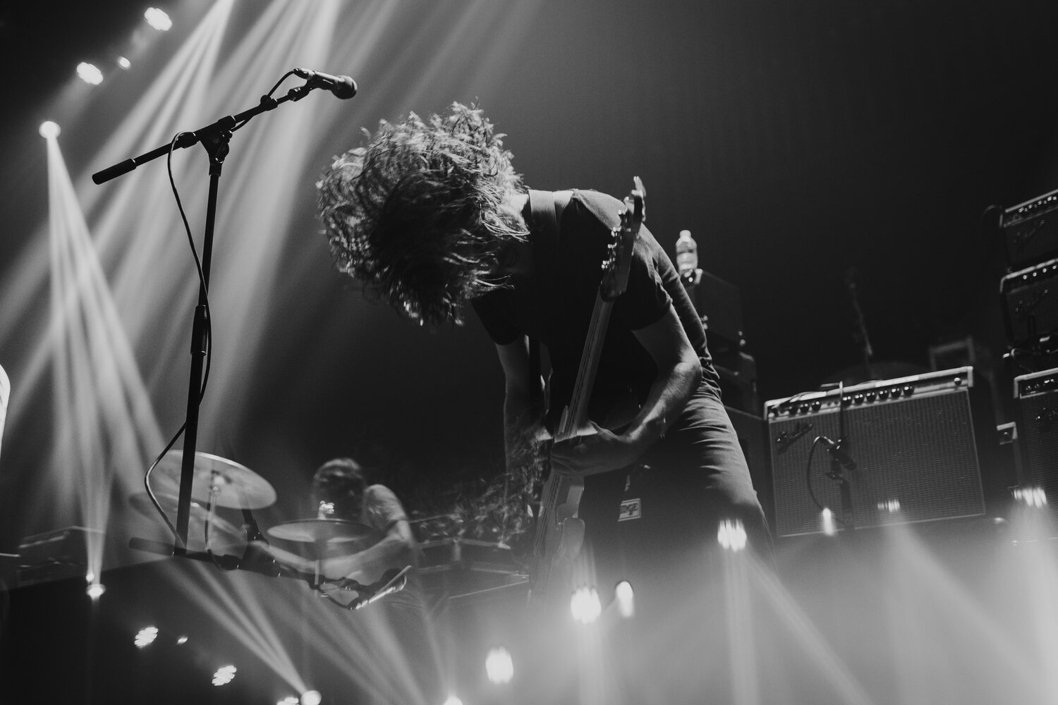 Black and white photo of a musician with long hair headbanging while playing guitar on stage, with stage lights and sound equipment in the background.