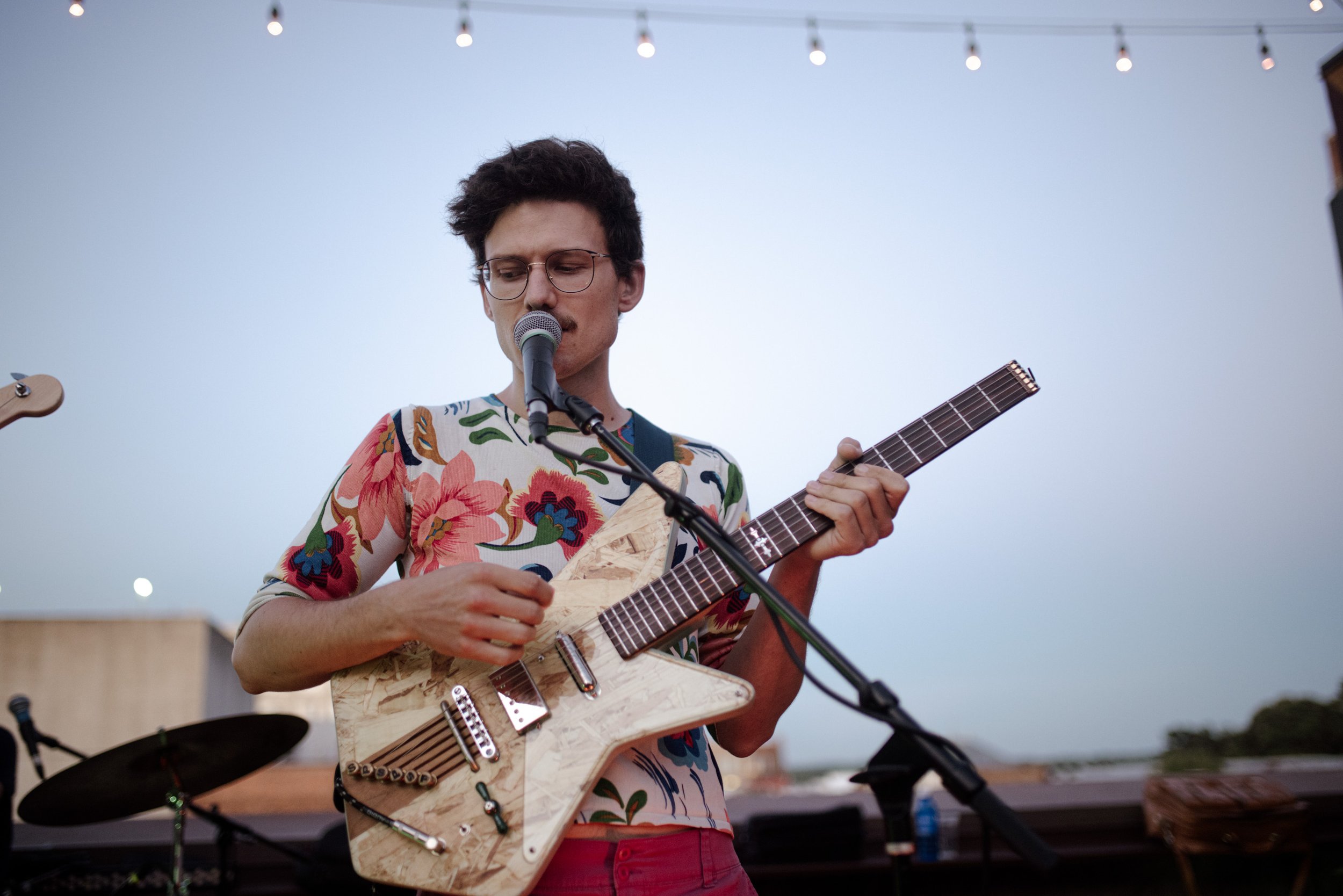 A young man with glasses and dark curly hair sings into a microphone while playing an electric guitar with a wooden finish at an outdoor event during dusk.