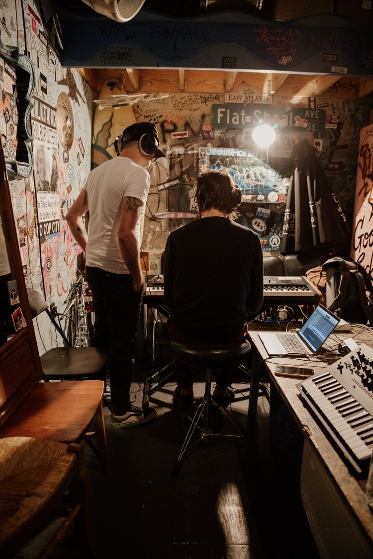 Two musicians in a graffiti-covered basement, one standing with headphones and a hat, the other seated at a keyboard, with a laptop and music equipment on a table nearby.