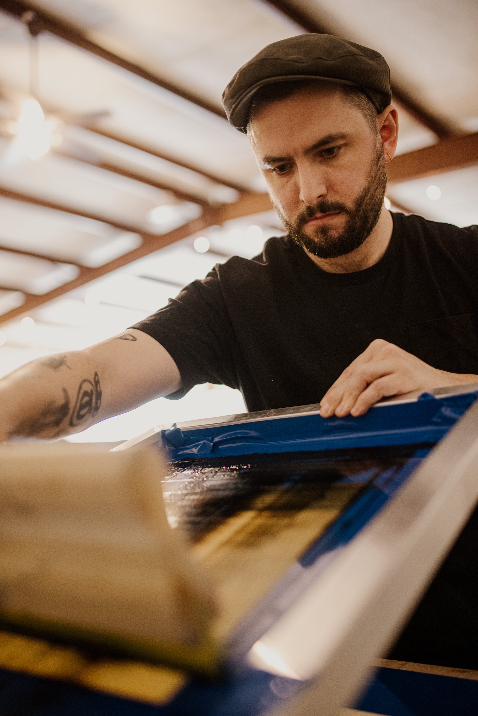 A man with tattoos on his arm and a beard, wearing a brown cap and black t-shirt, appears to be examining or working on a framed piece of artwork or photograph on a flat surface indoors.