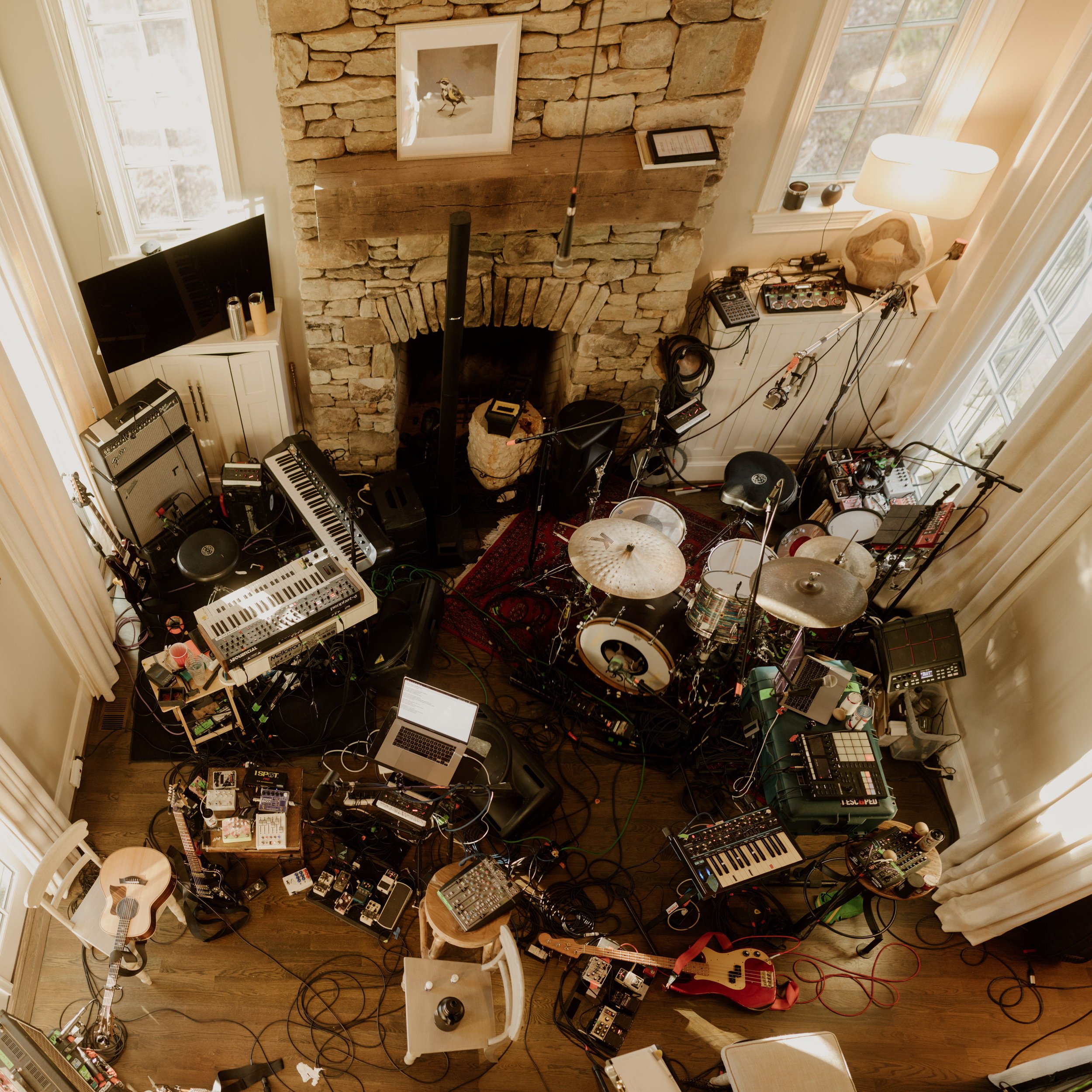 A room set up as a music studio with a recording drum kit, electronic keyboards, mixers, laptops, guitars, microphones, and audio equipment, arranged in a cozy space with a stone fireplace, large windows, and curtains.