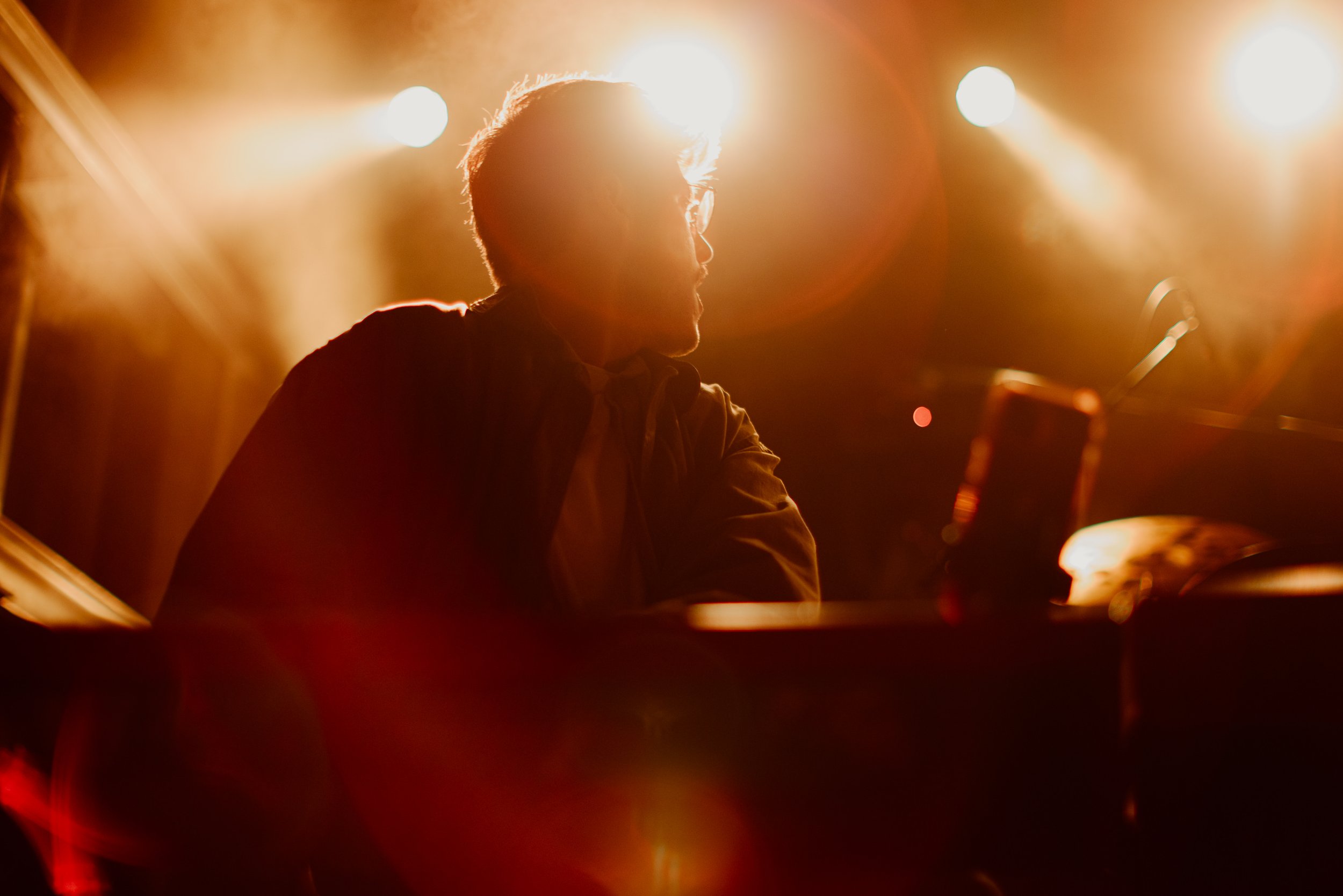 Silhouette of a man sitting at a piano or keyboard on stage, illuminated by bright stage lights behind him, creating a backlit effect.