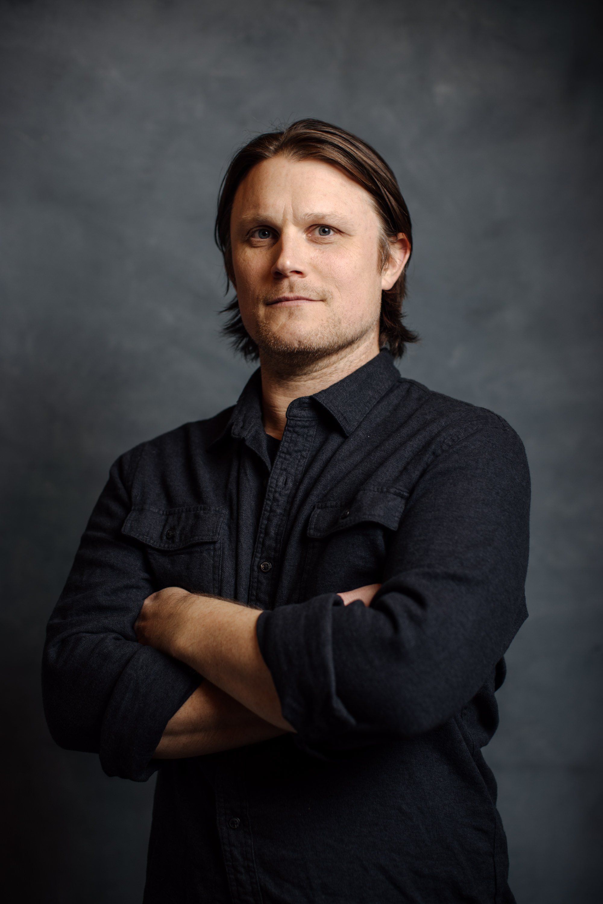 A man with light skin and brown hair, wearing a black button-up shirt, standing with crossed arms against a dark gray background.