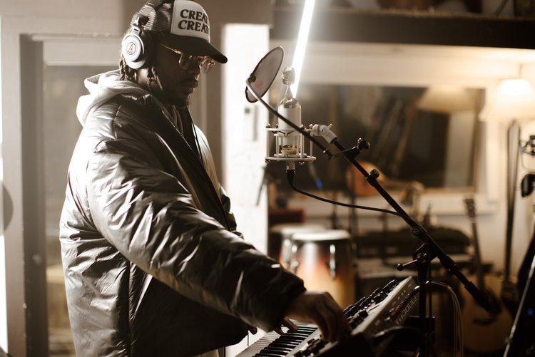 Person wearing headphones, glasses, fur-lined jacket, and a cap with the word 'CREW', playing a keyboard in a music studio with a microphone overhead.