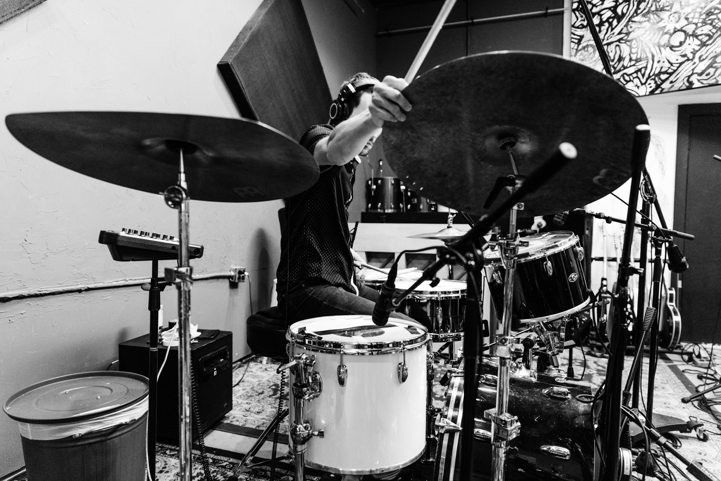 Person playing drums in a recording studio, surrounded by microphones and musical equipment, with a wall art piece in the background.