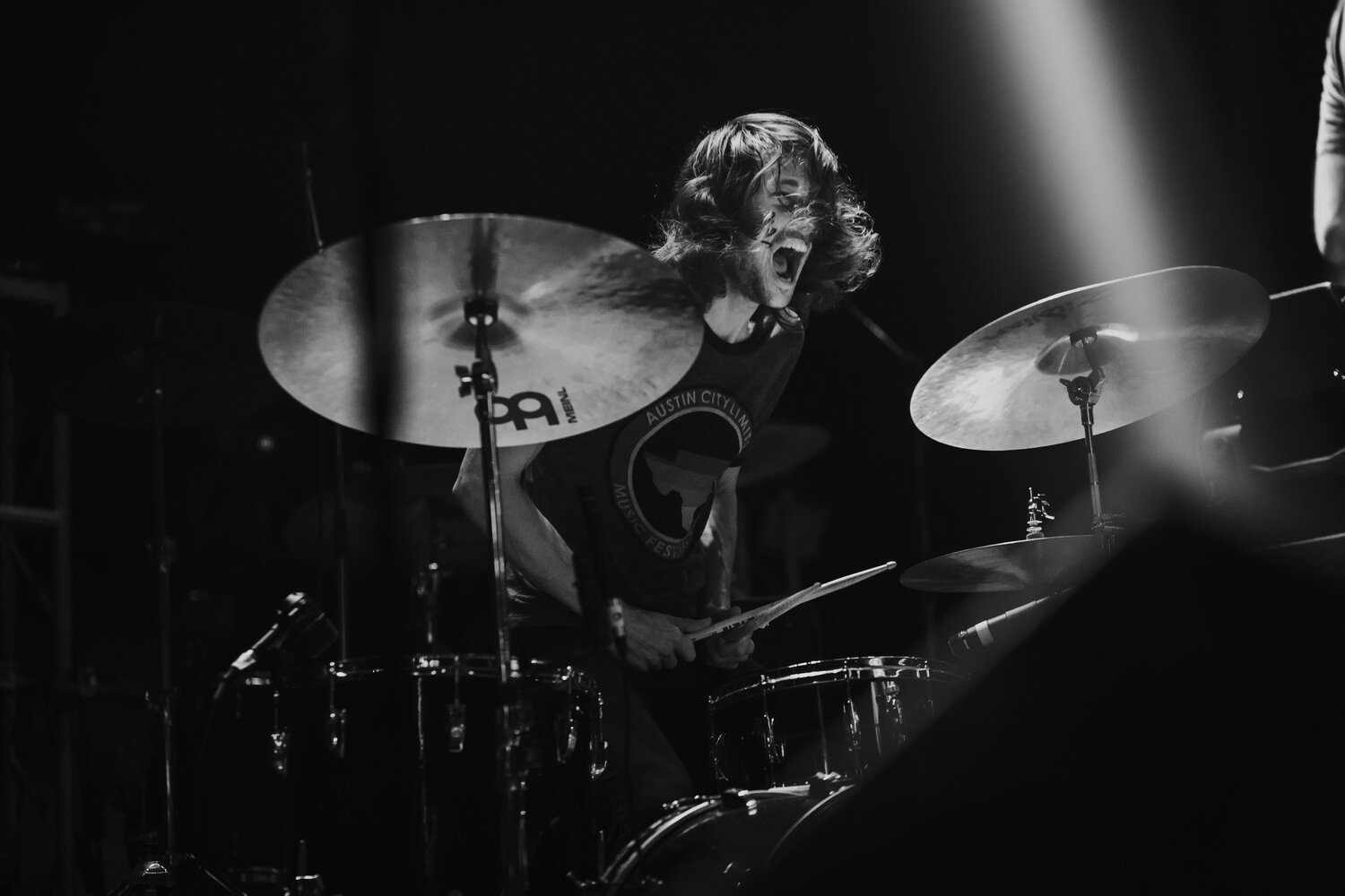 Black and white photo of a drummer passionately playing a drum set on stage, with a wide-open mouth and curly hair.