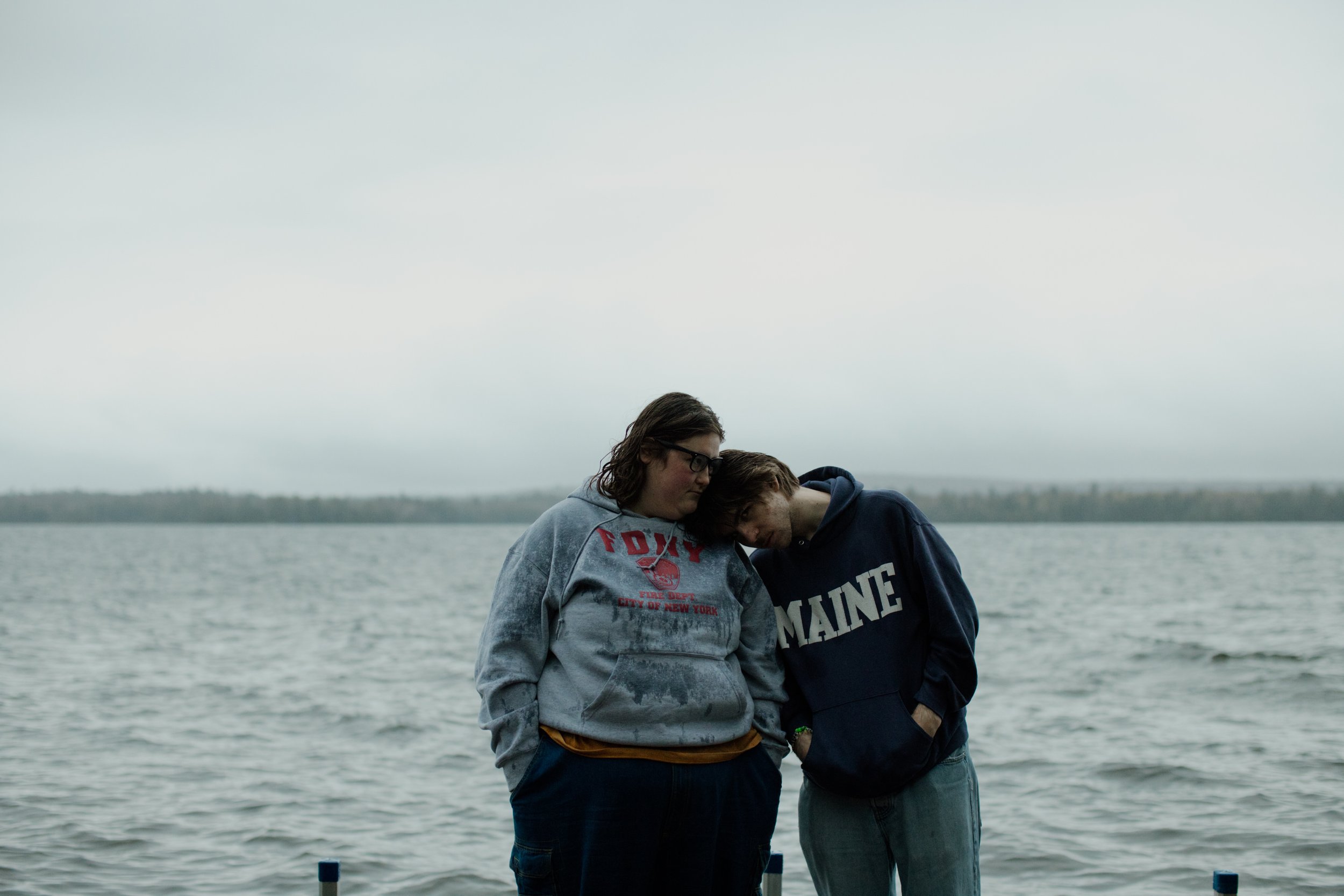 Two young people stand close together near a body of water, with their heads touching and eyes closed, appearing contemplative or comforting each other on a cloudy day.