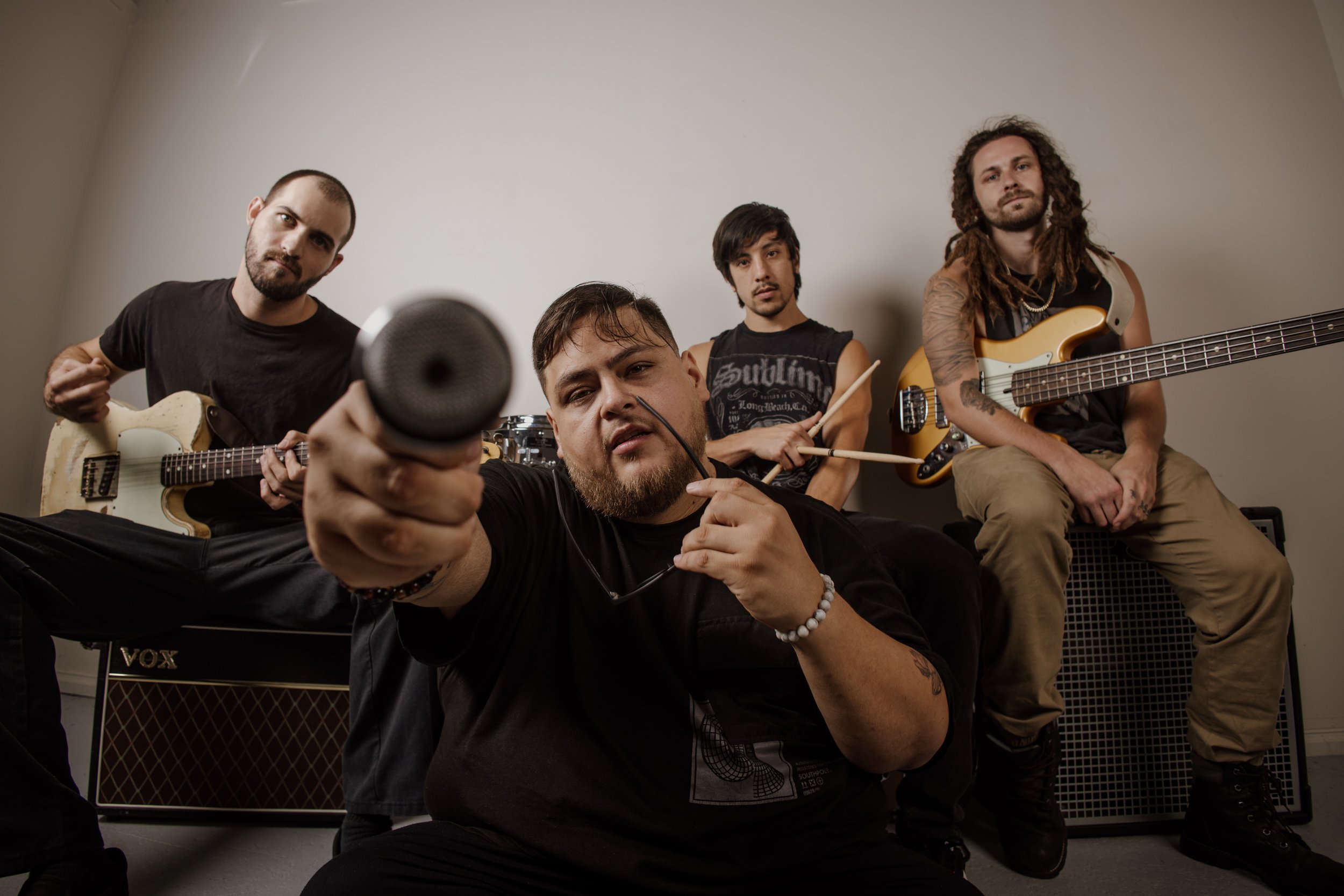 Four male musicians posing together in a room, with one holding a microphone, two holding guitars, and one with drumsticks, all looking at the camera.