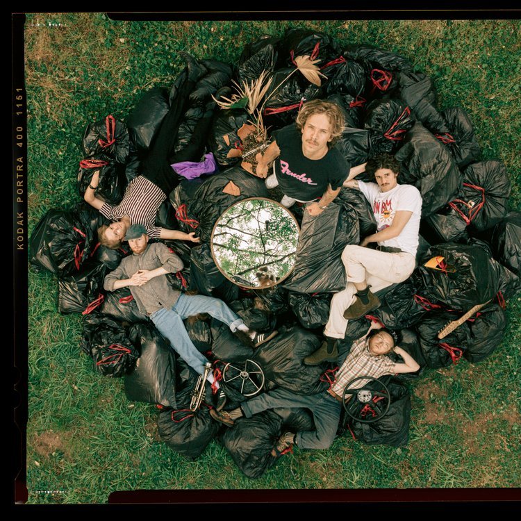 Seven people lying on black garbage bags arranged in a circle on a grassy field, viewed from above, with a reflective circular mirror showing tree branches in the center.