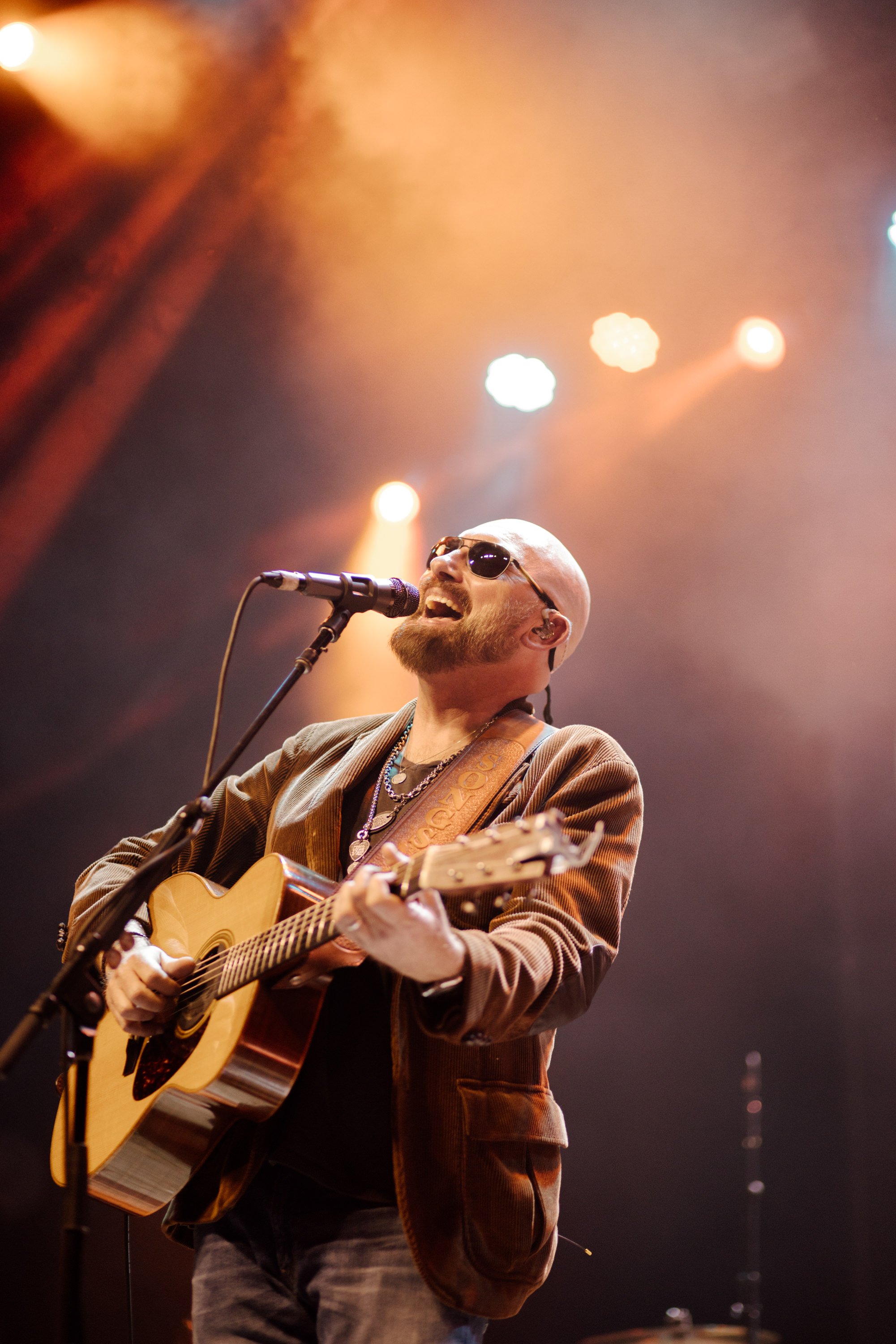 A man with a beard and sunglasses playing an acoustic guitar and singing into a microphone during a live music performance on stage, with orange stage lights in the background.