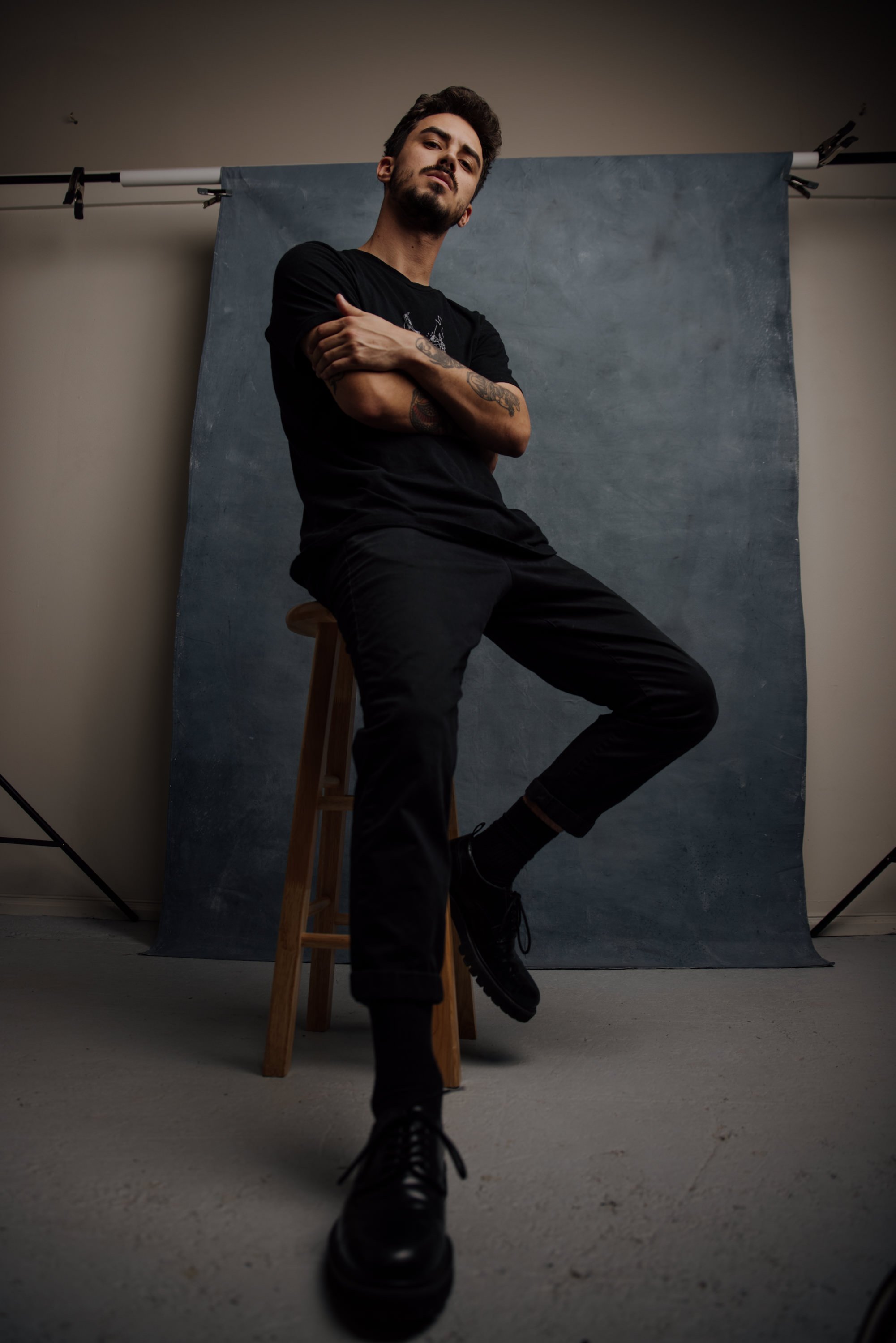 Young man with dark hair and beard sitting on a wooden stool in front of a gray backdrop, wearing a black t-shirt, black pants, and black shoes, looking confidently at the camera with arms crossed.