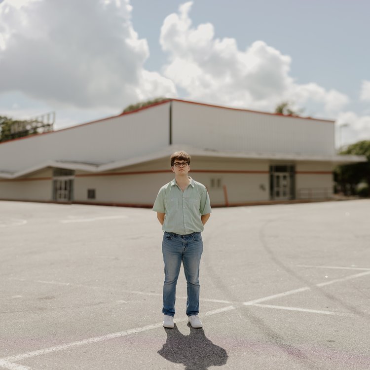Elijah Johnston A person standing alone in an empty parking lot in front of a large white building with a sloped roof, under a cloudy sky.