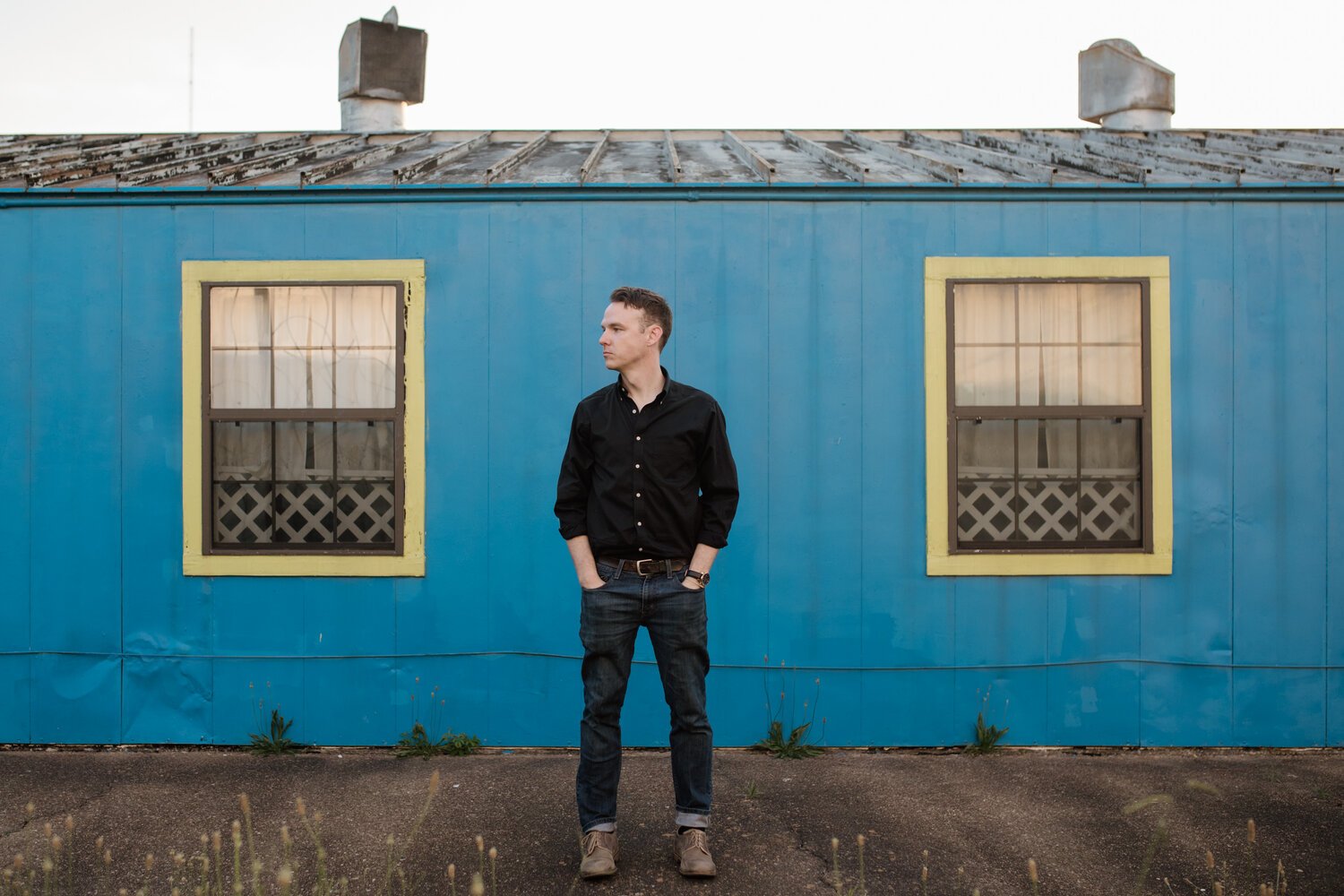 A man standing with hands in pockets in front of a blue building with two windows with yellow borders.