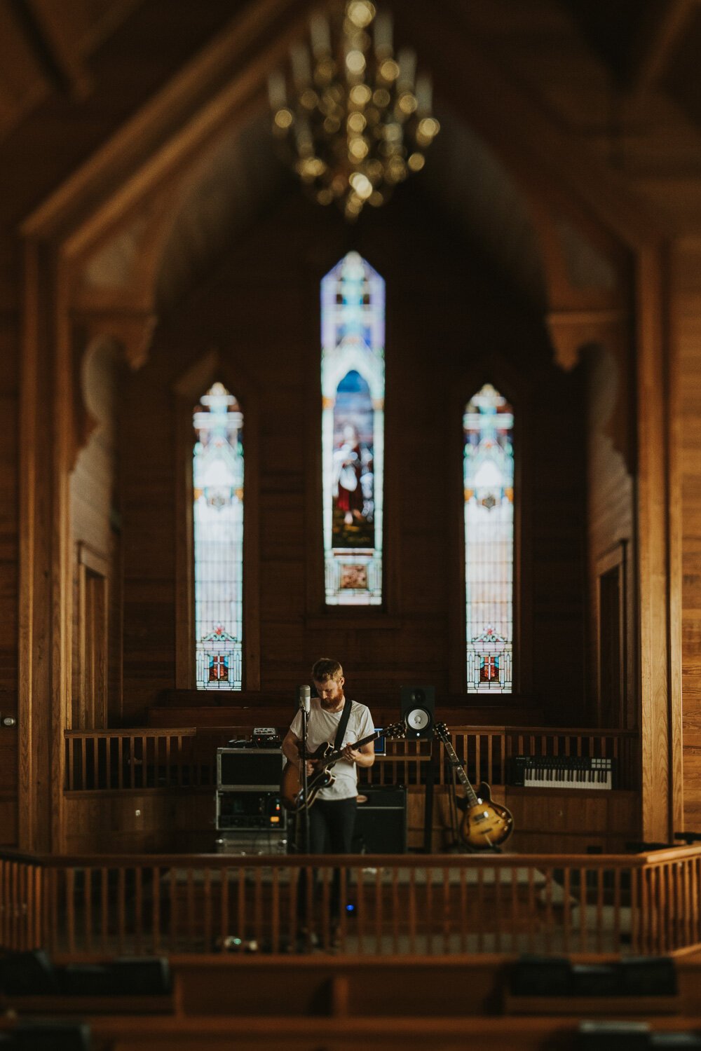 A man playing guitar on stage inside a wooden church with stained glass windows in the background.