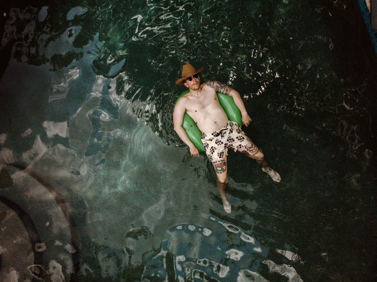 Man wearing a cowboy hat and sunglasses, sitting in a green inflatable float in a pool, with tattoos on his arms and legs, wearing patterned swim trunks.