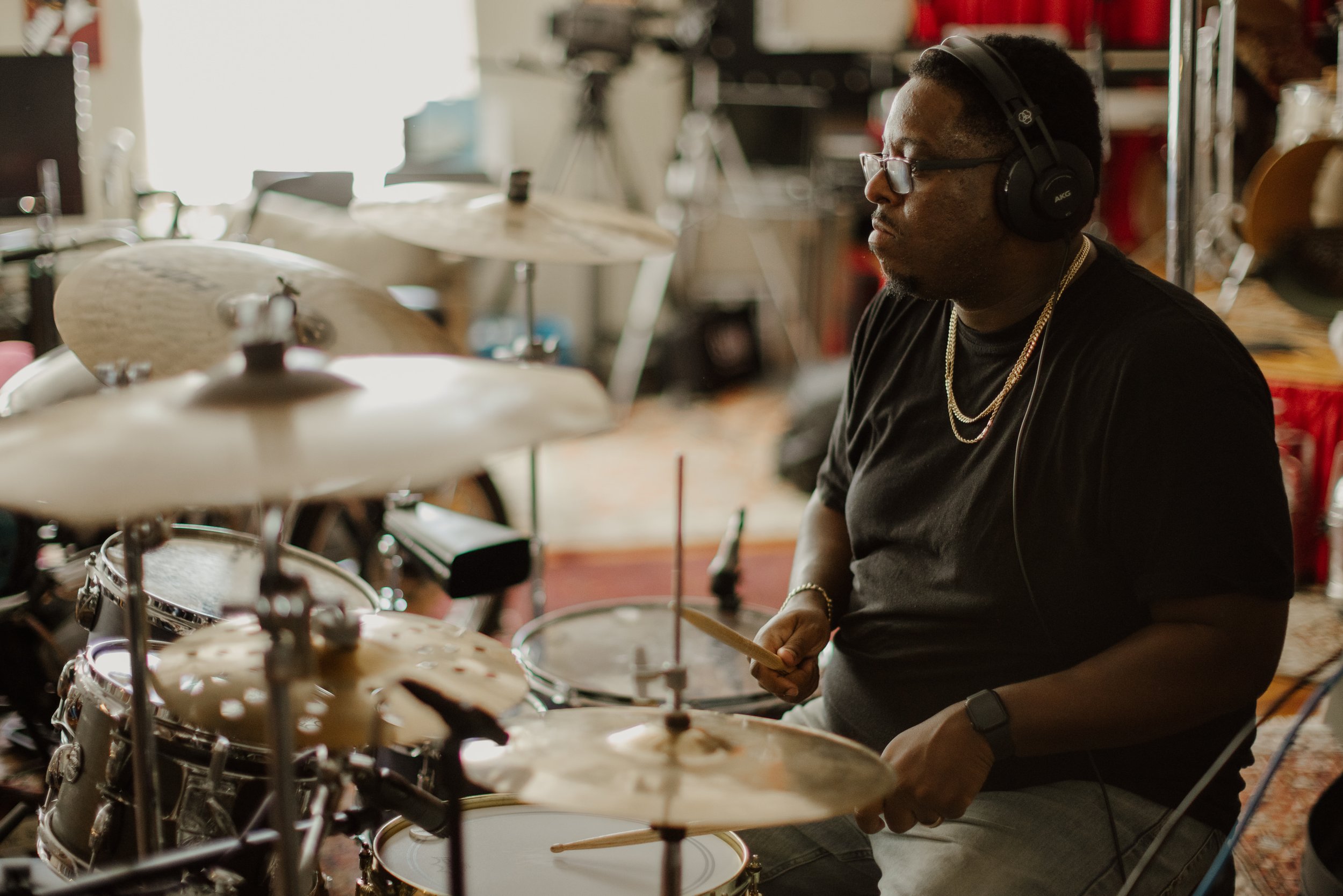 A man wearing glasses, headphones, and a black t-shirt playing a drum set in a music studio.