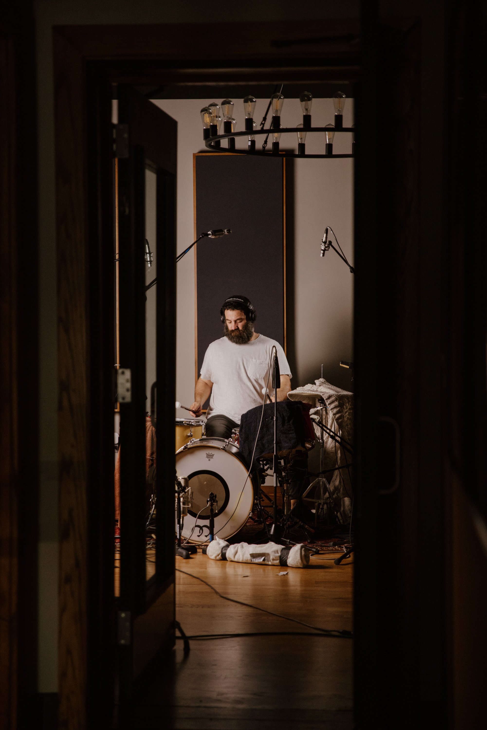 A bearded man with headphones playing drums in a recording studio through a doorway, with microphones and a chandelier overhead.
