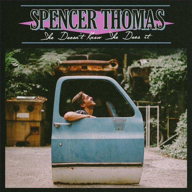 A man sitting inside a car door with the window rolled down, looking up with a relaxed expression, with two abandoned cars in the background and greenery around, on what appears to be a driveway or lot. The top of the image has the text "Spencer Thom