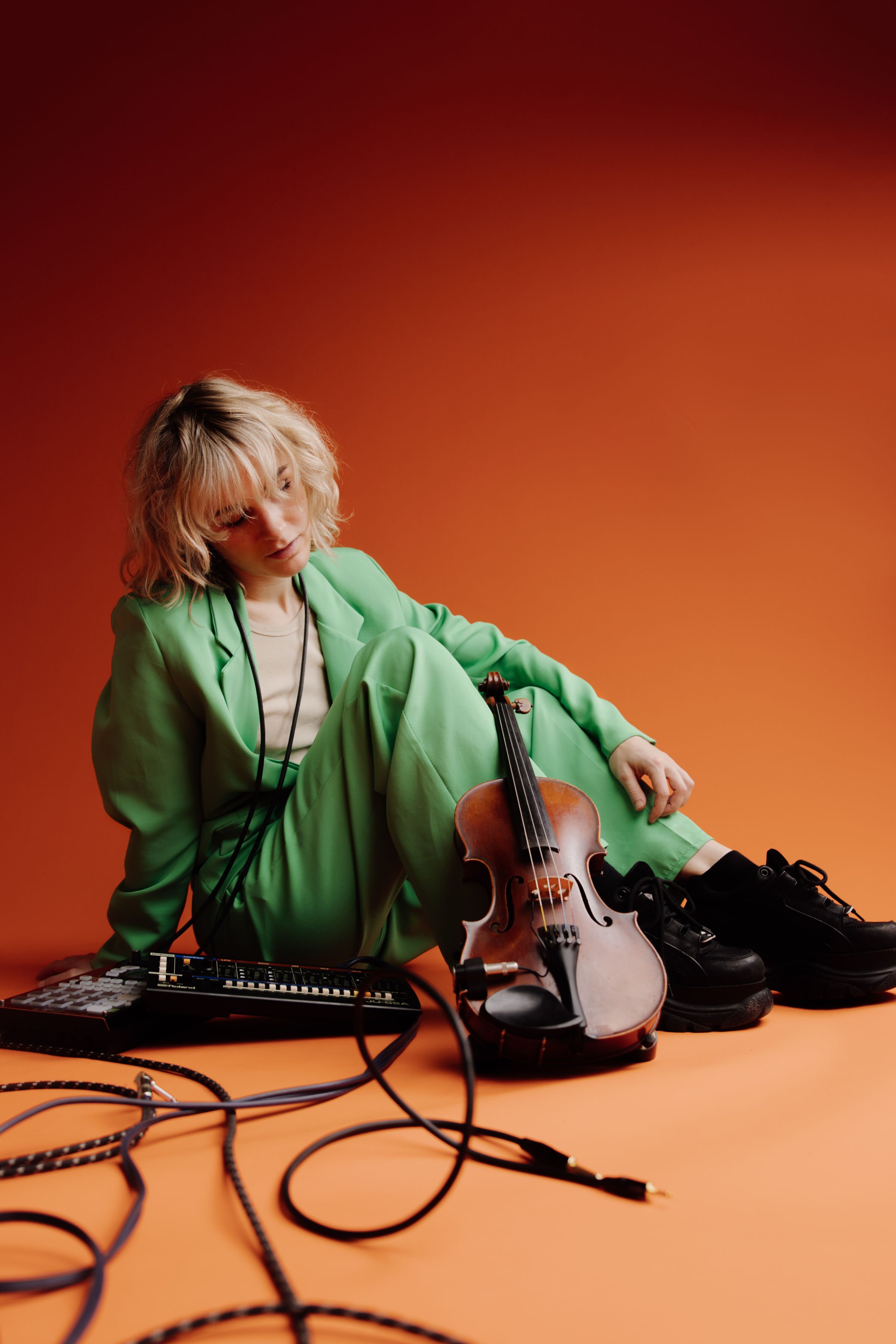 Young woman with blonde curly hair, dressed in a green suit, sitting on an orange floor with a violin, a music controller, and various cables, against an orange backdrop.