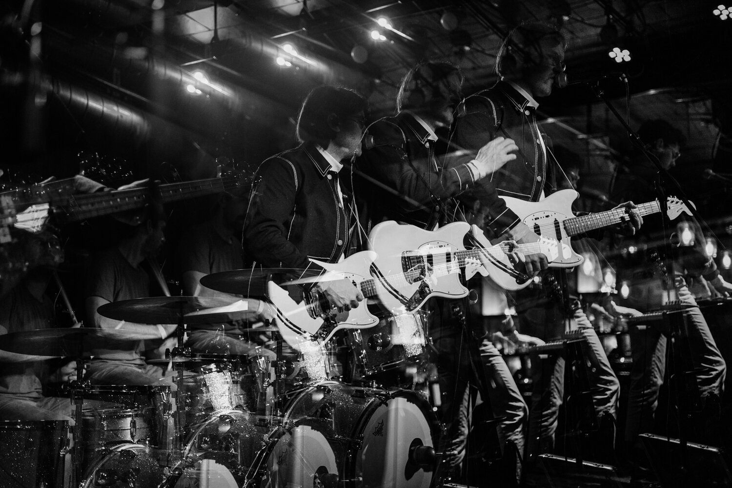 Black and white photo of a live band performance with multiple musicians playing electric guitars and drums, stage lights creating a moody atmosphere.