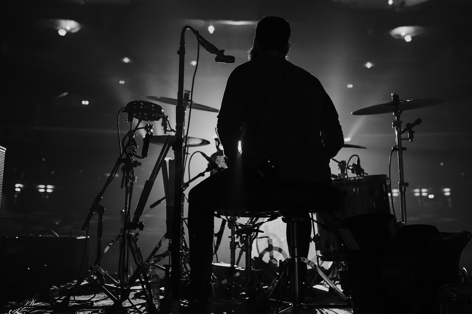 Silhouette of a drummer on stage with drums and microphones, backlit creating a dramatic black and white scene.