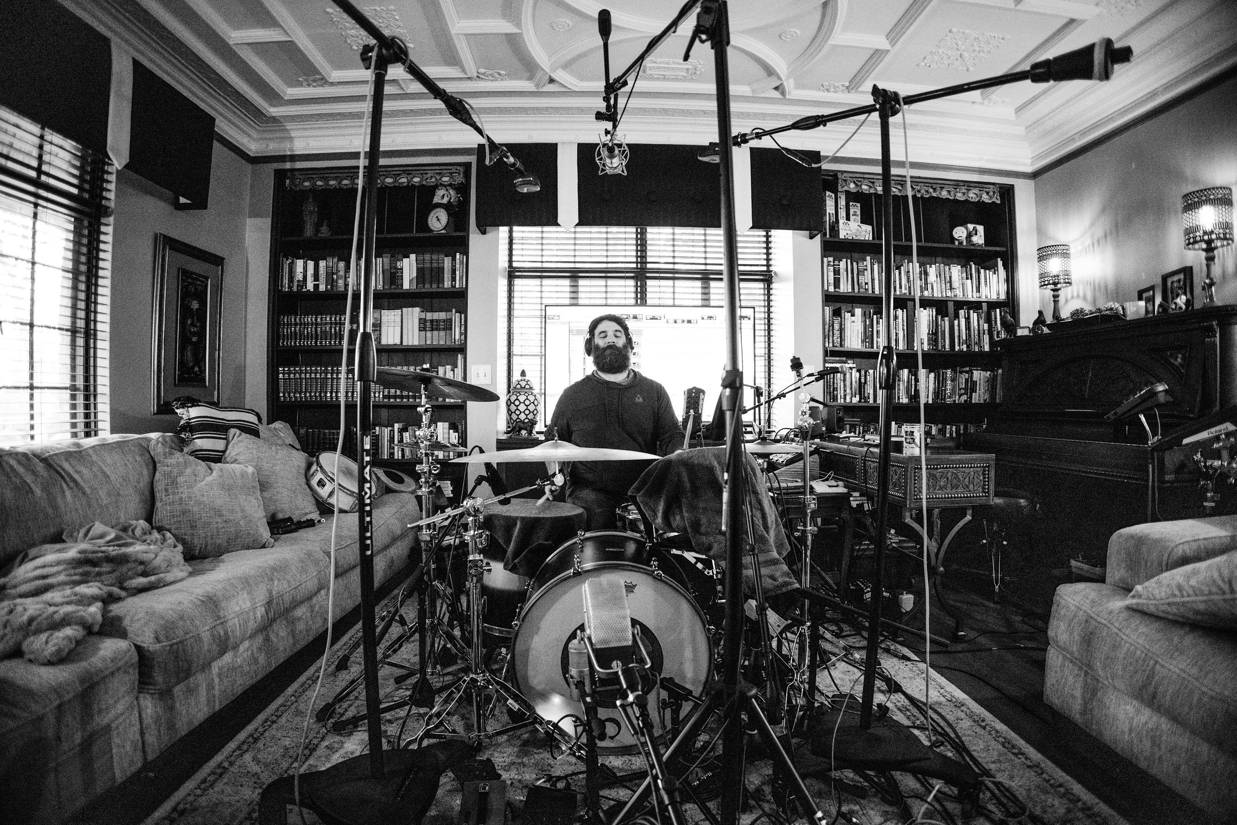 A man sitting behind a drum set in a living room with bookshelves, a piano, and recording microphones overhead.