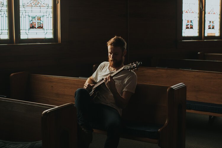 A man with a beard playing an acoustic guitar in a church pew, with stained glass windows in the background.