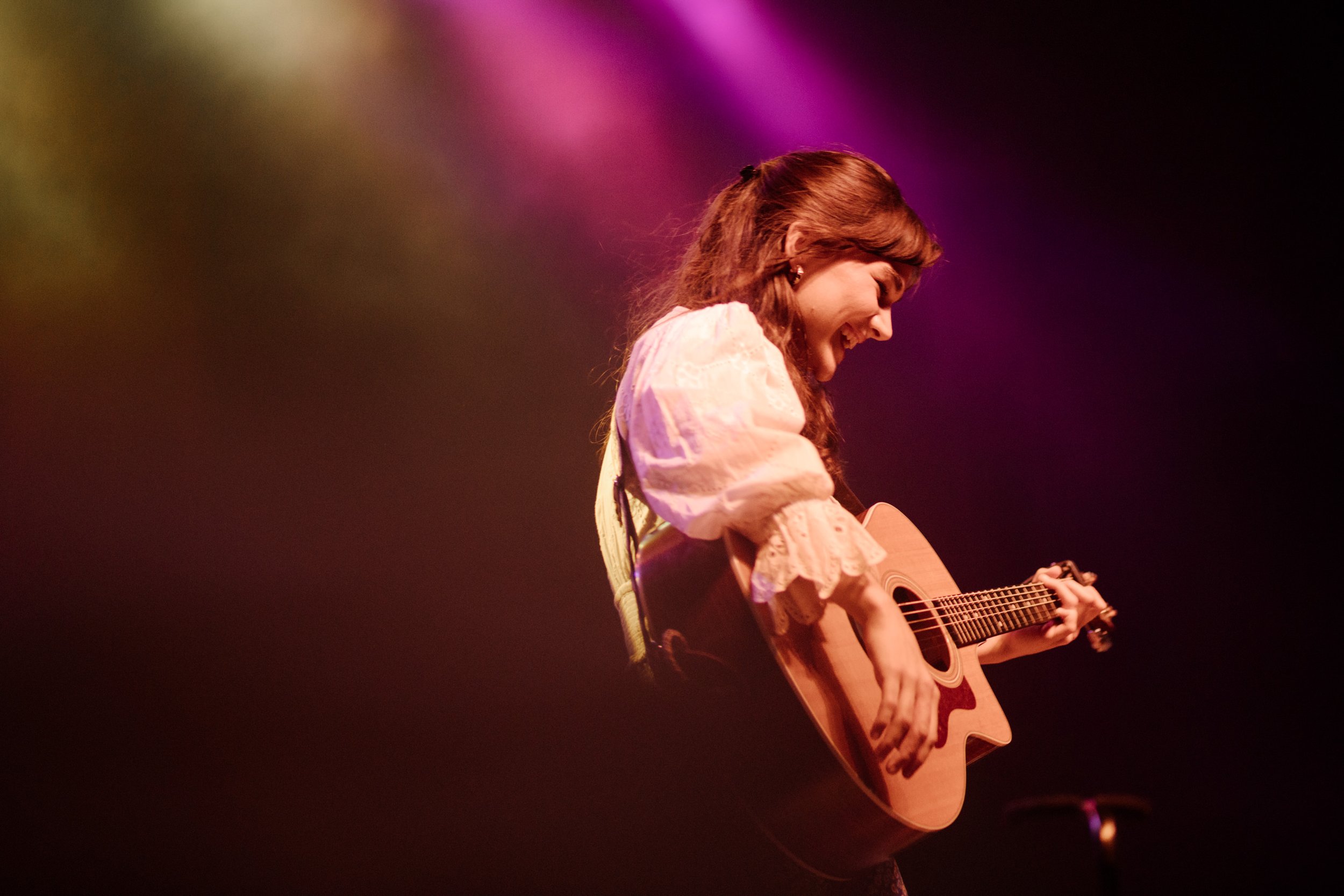 A young woman with long curly hair, wearing a white blouse with puffed sleeves, smiling and playing an acoustic guitar on stage, illuminated by colorful stage lights.