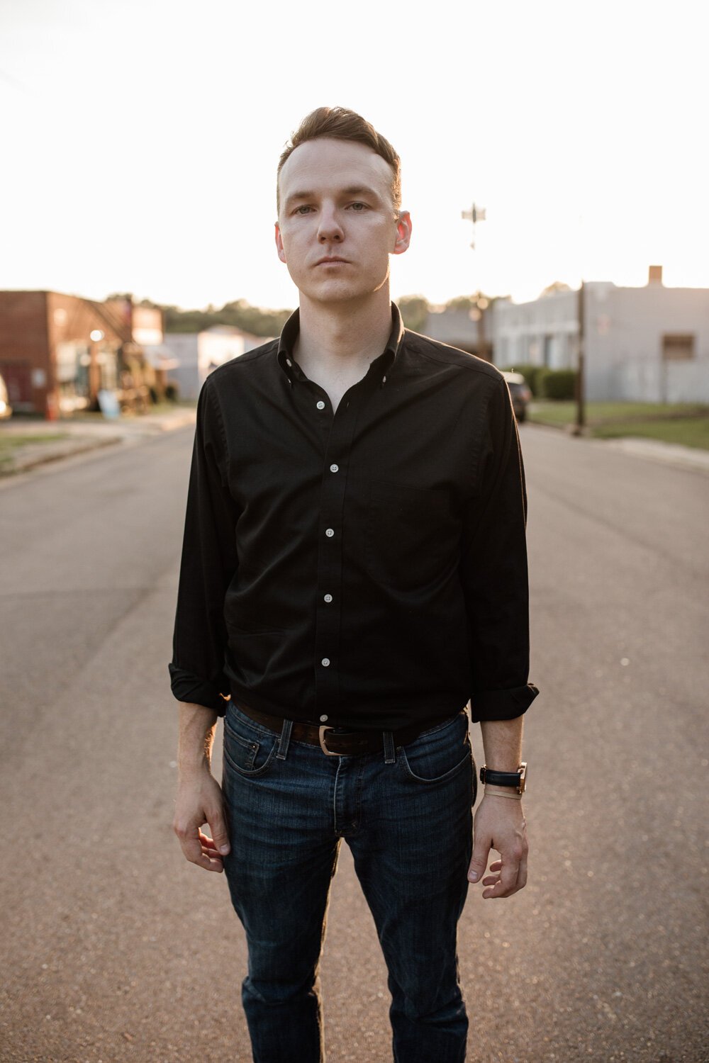 A man with short brown hair, wearing a black button-down shirt and dark jeans, stands in the middle of a quiet street during sunset, with buildings and a clear sky in the background.
