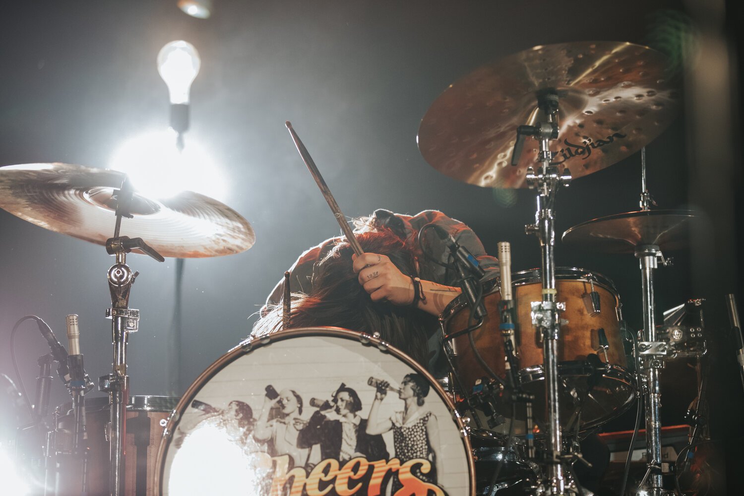 Female drummer with long hair, head down, holding drumsticks, surrounded by cymbals and drums on stage during a concert.