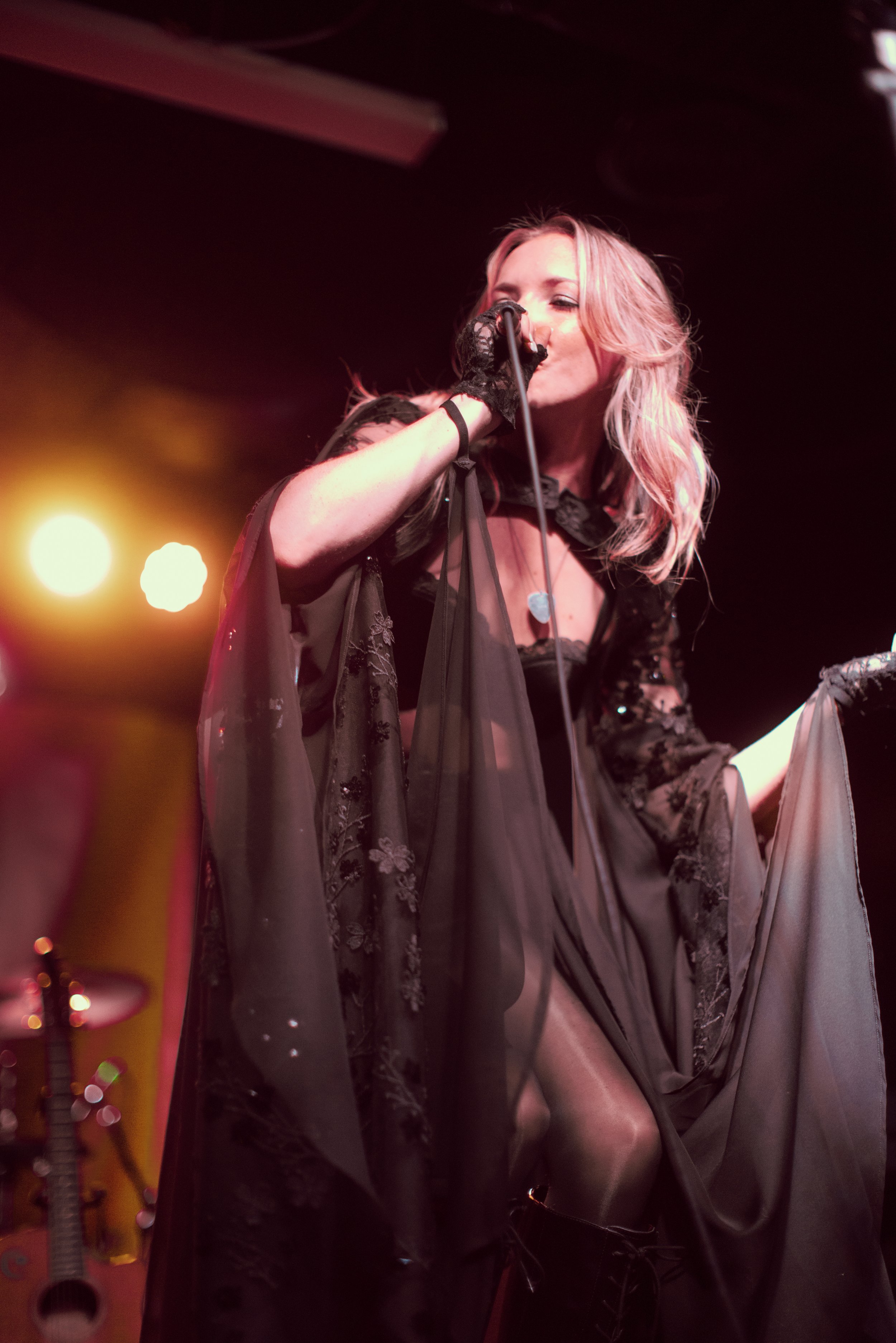 Female singer on stage wearing a sheer black lace dress, holding a microphone and singing. Blonde hair, stage lights in the background.