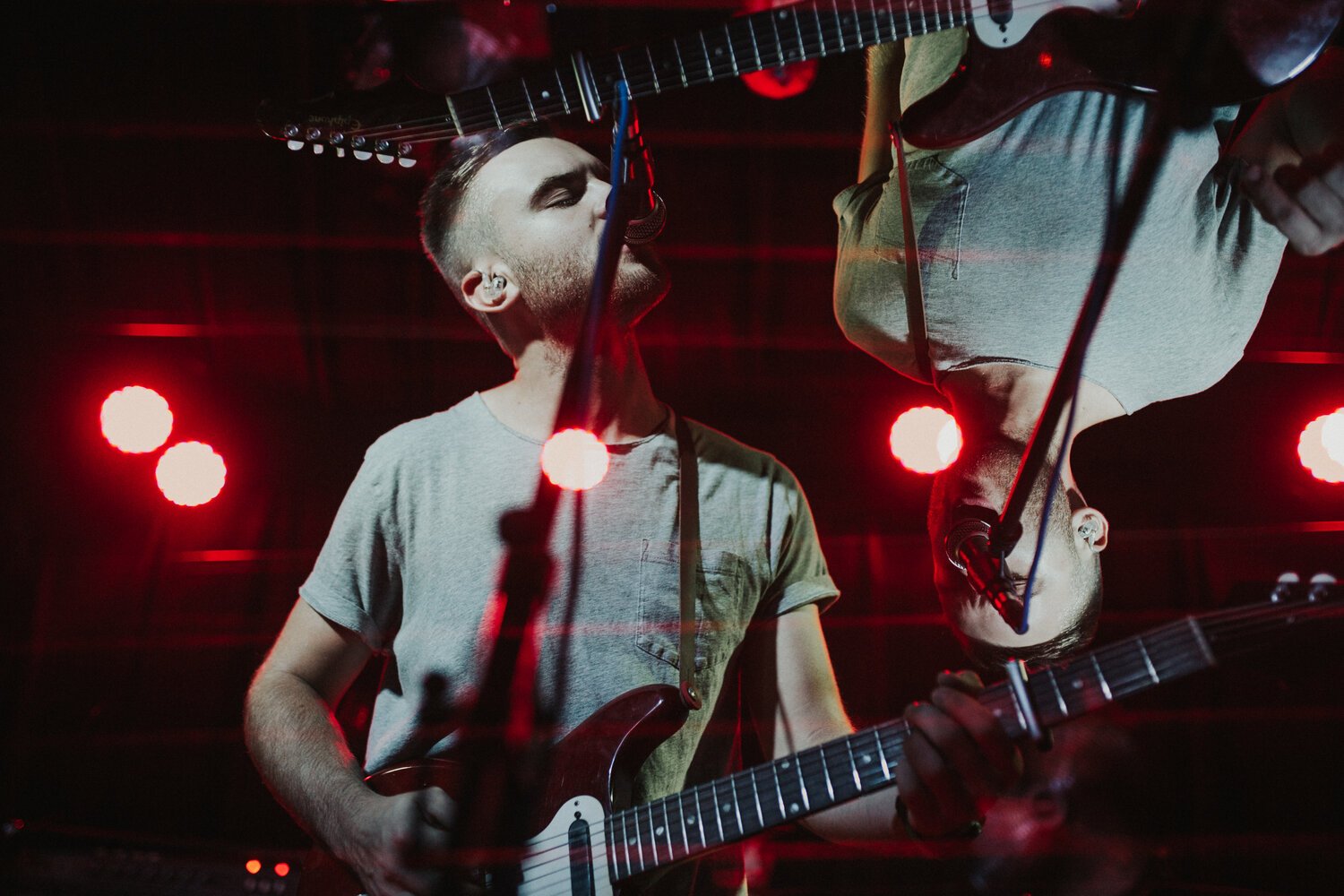 A musician playing an electric guitar on stage under red stage lights, with a reflective surface showing part of his face and top of his head.