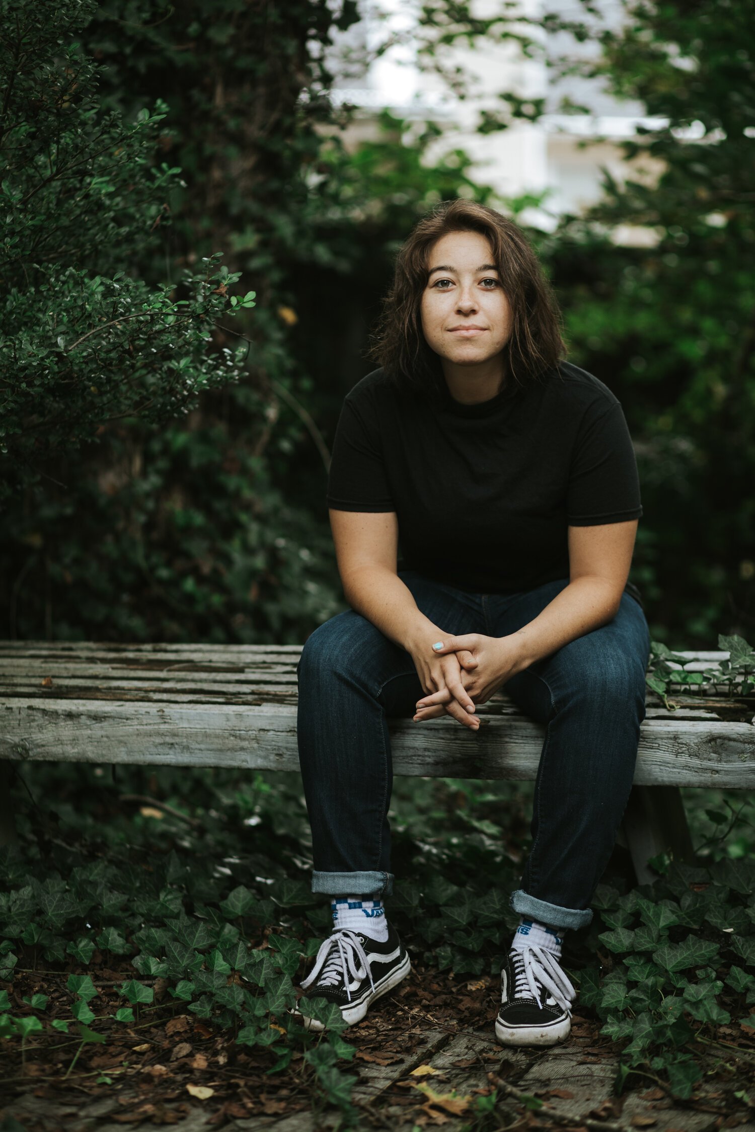 Woman sitting on a weathered wooden bench outdoors, surrounded by green foliage, wearing a black t-shirt, dark jeans, and black and white sneakers.