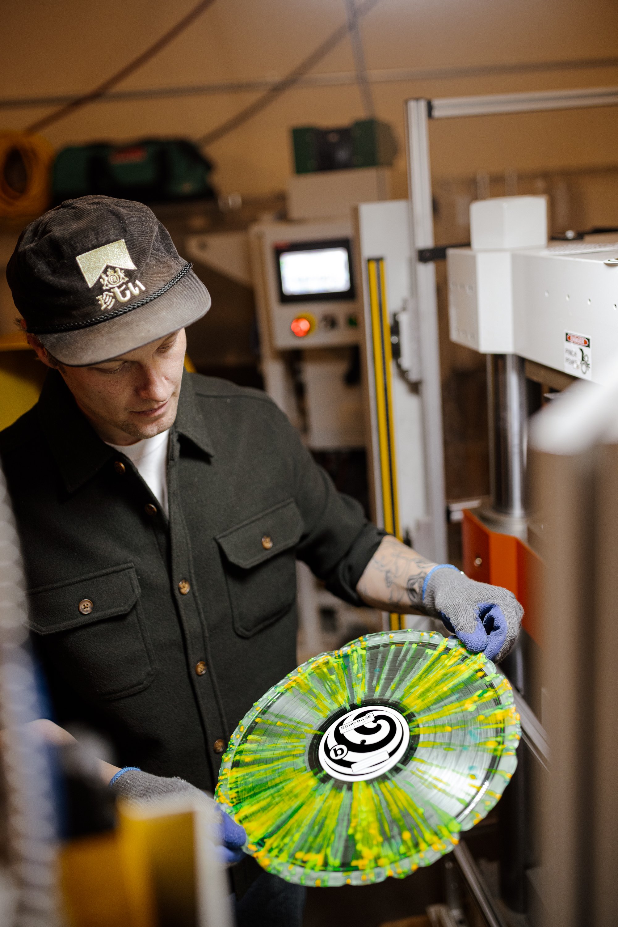 Echo Base Athens Ga A person wearing a black cap and black shirt holding a colorful vinyl record in a workshop setting.