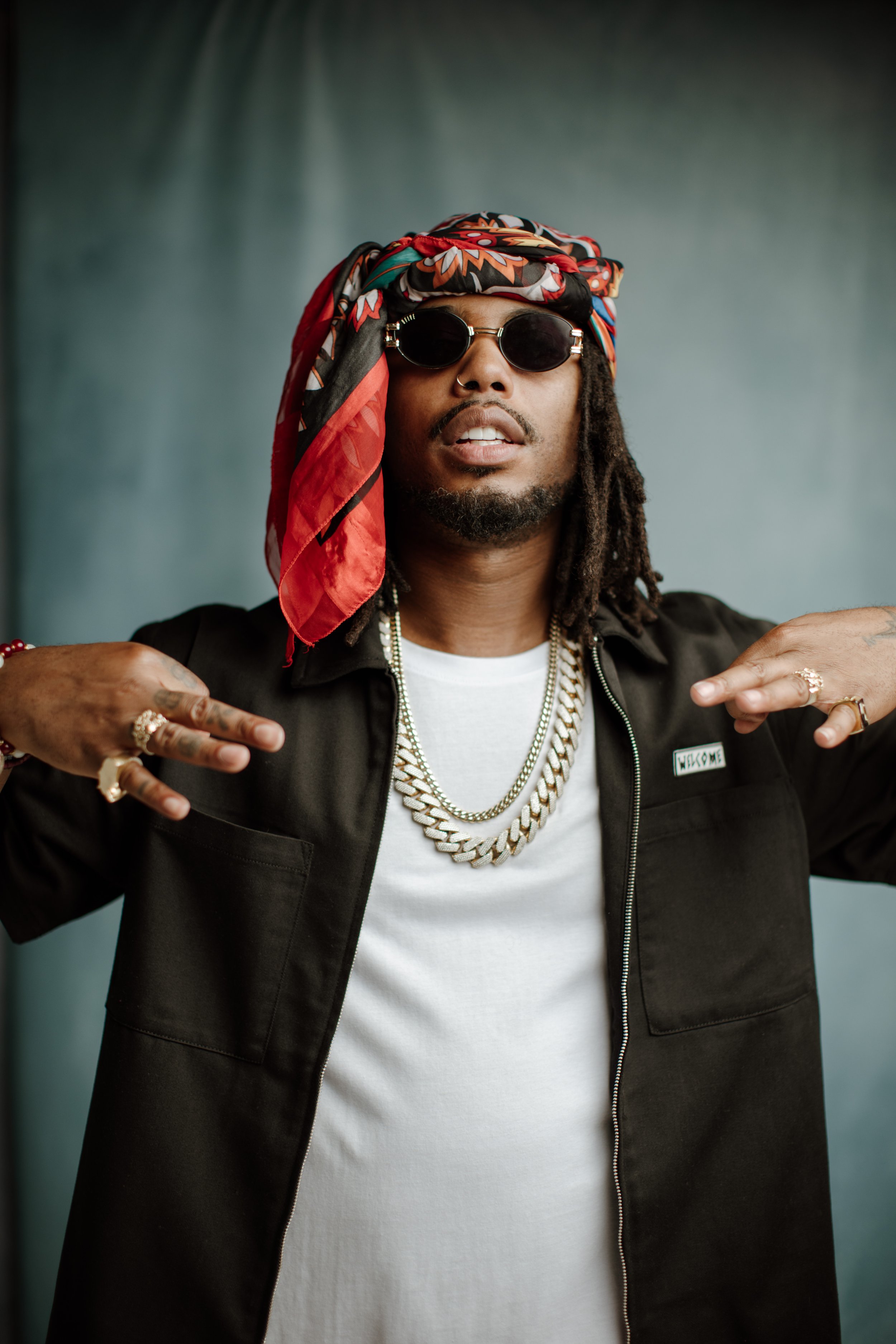 A man with dreadlocks wearing a colorful headscarf, sunglasses, a black jacket, a white shirt, and multiple gold chains, posing with his hands near his shoulders.