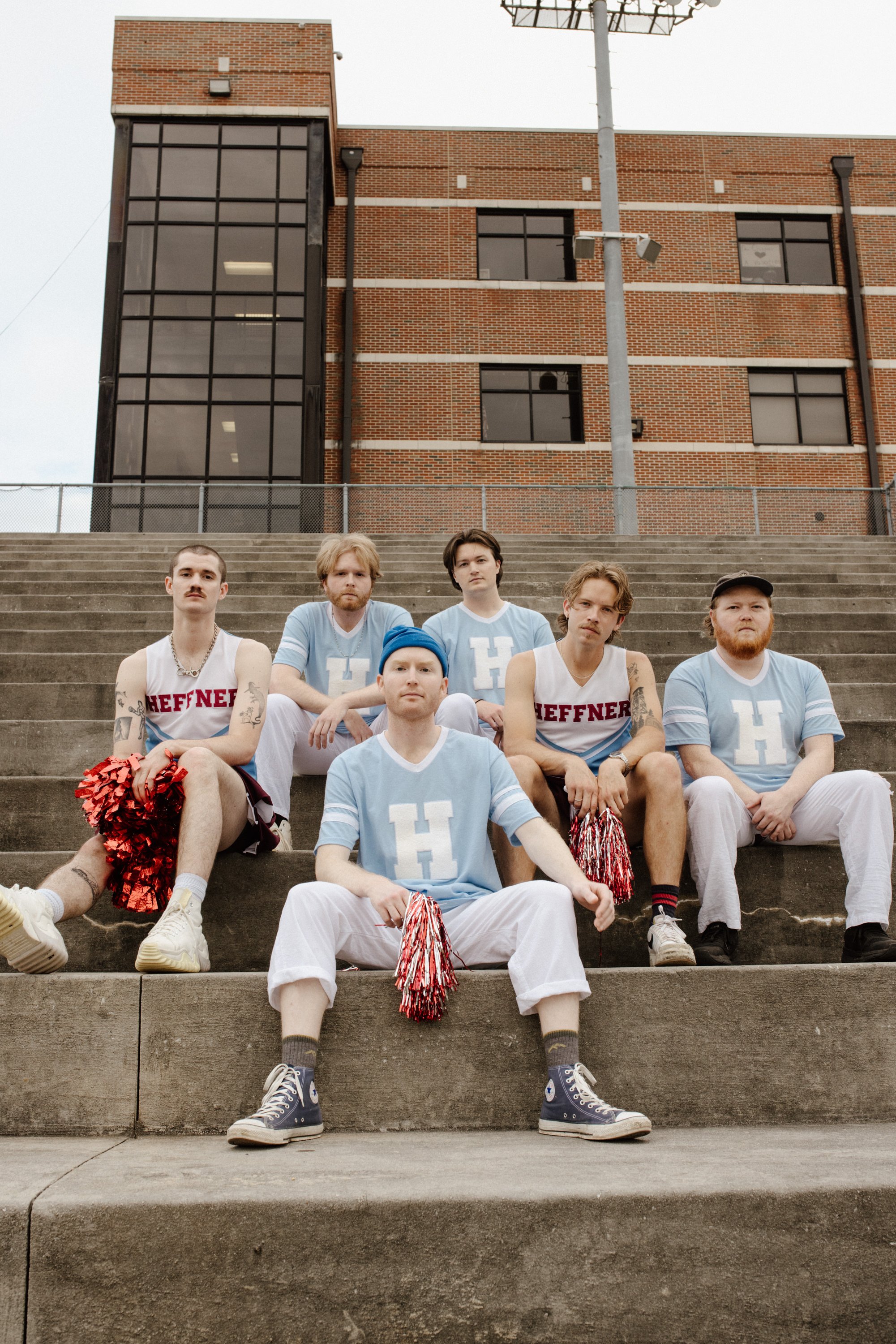 Group of six young men sitting on outdoor stadium steps dressed in cheerleading and sports uniforms, with a brick building and utility poles in the background.