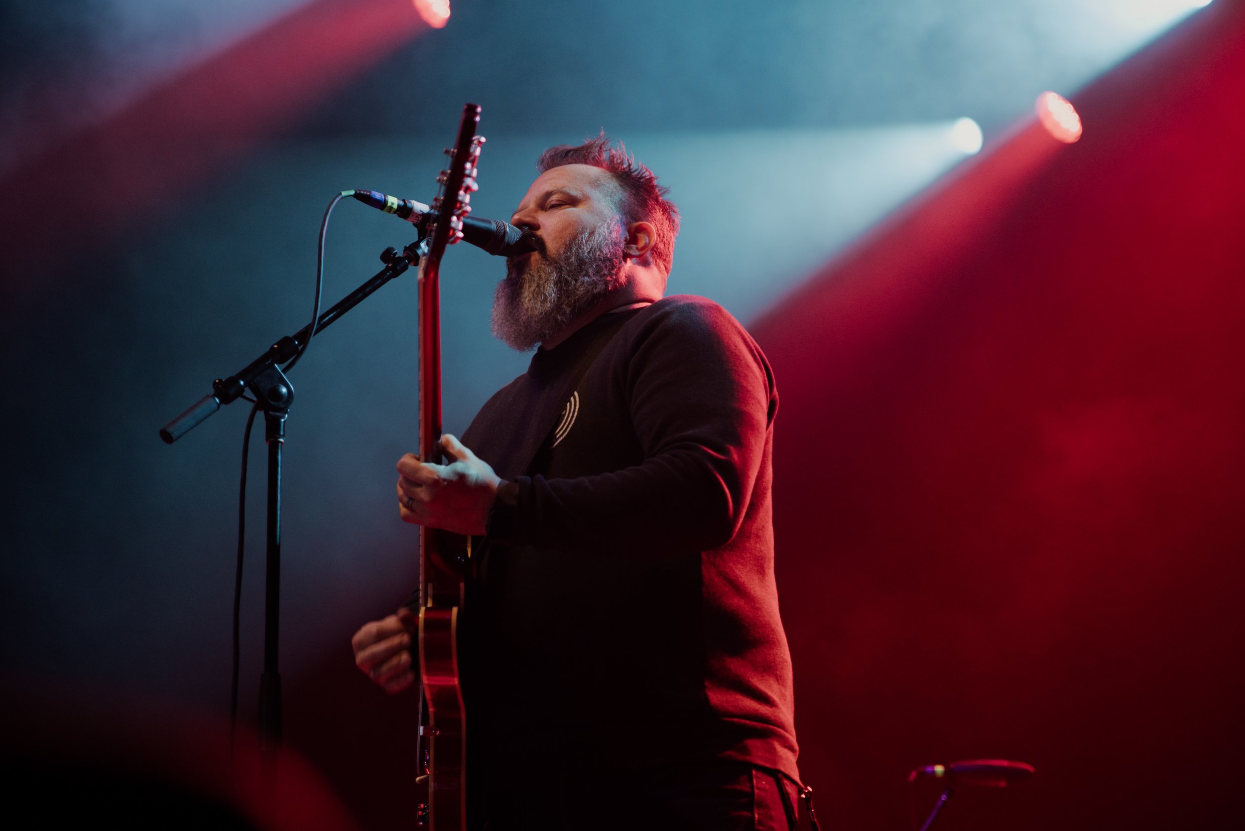 Man with a beard singing into a microphone while playing an electric guitar on stage with red and blue lighting.